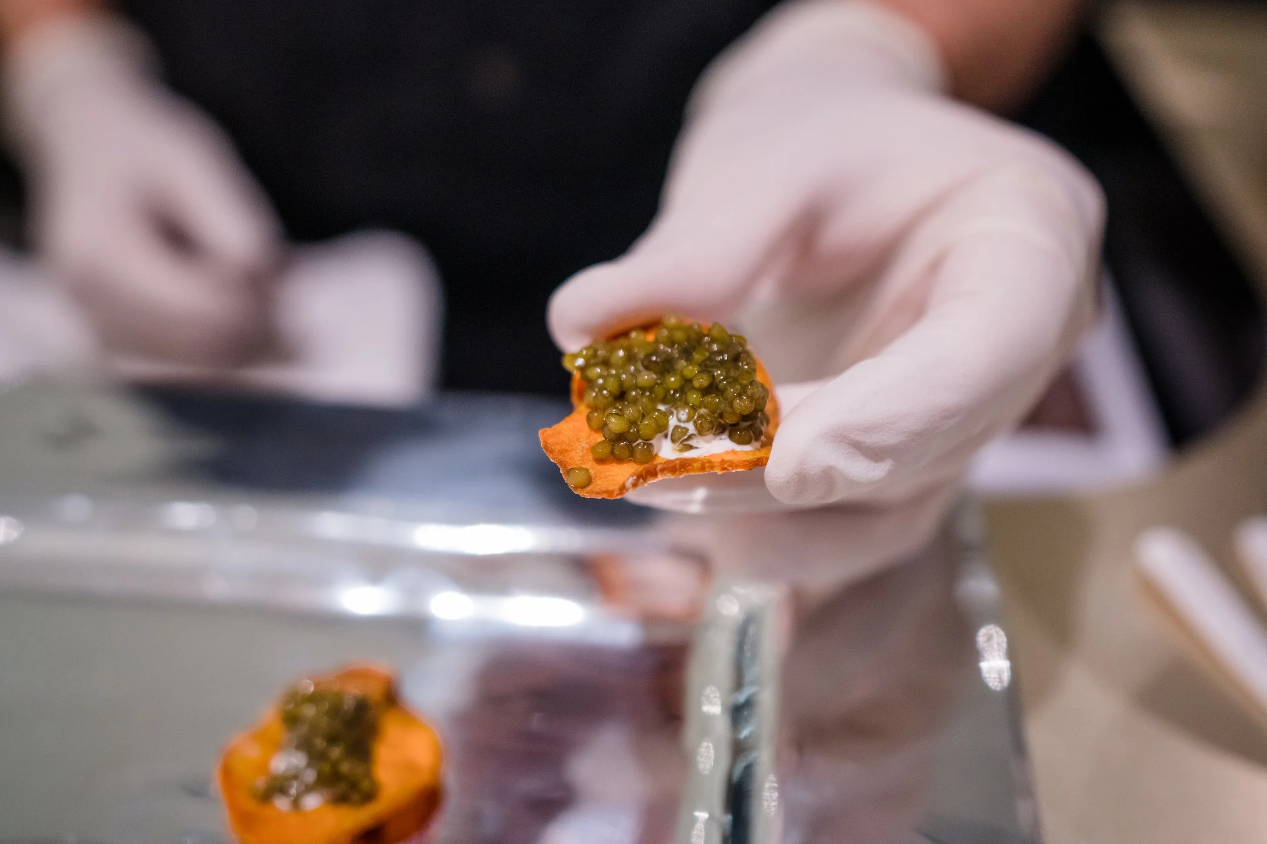 Close-up of a gloved hand holding an orange spice decoration topped with green caviar-like pearls, with a similar decoration resting on a shiny metal surface.