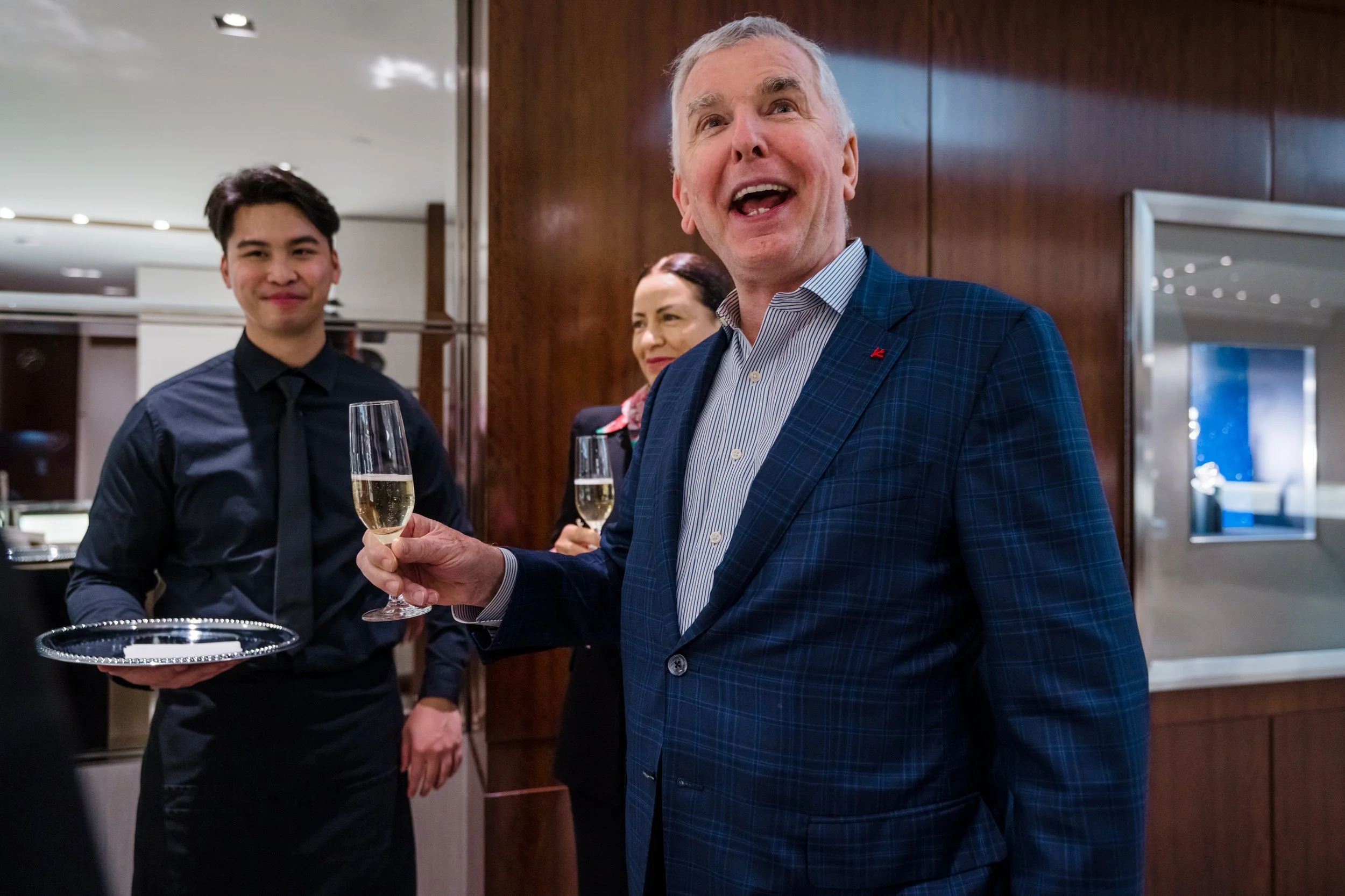 A man in a blue checked suit holding a glass of champagne at a celebration event with two people in the background.