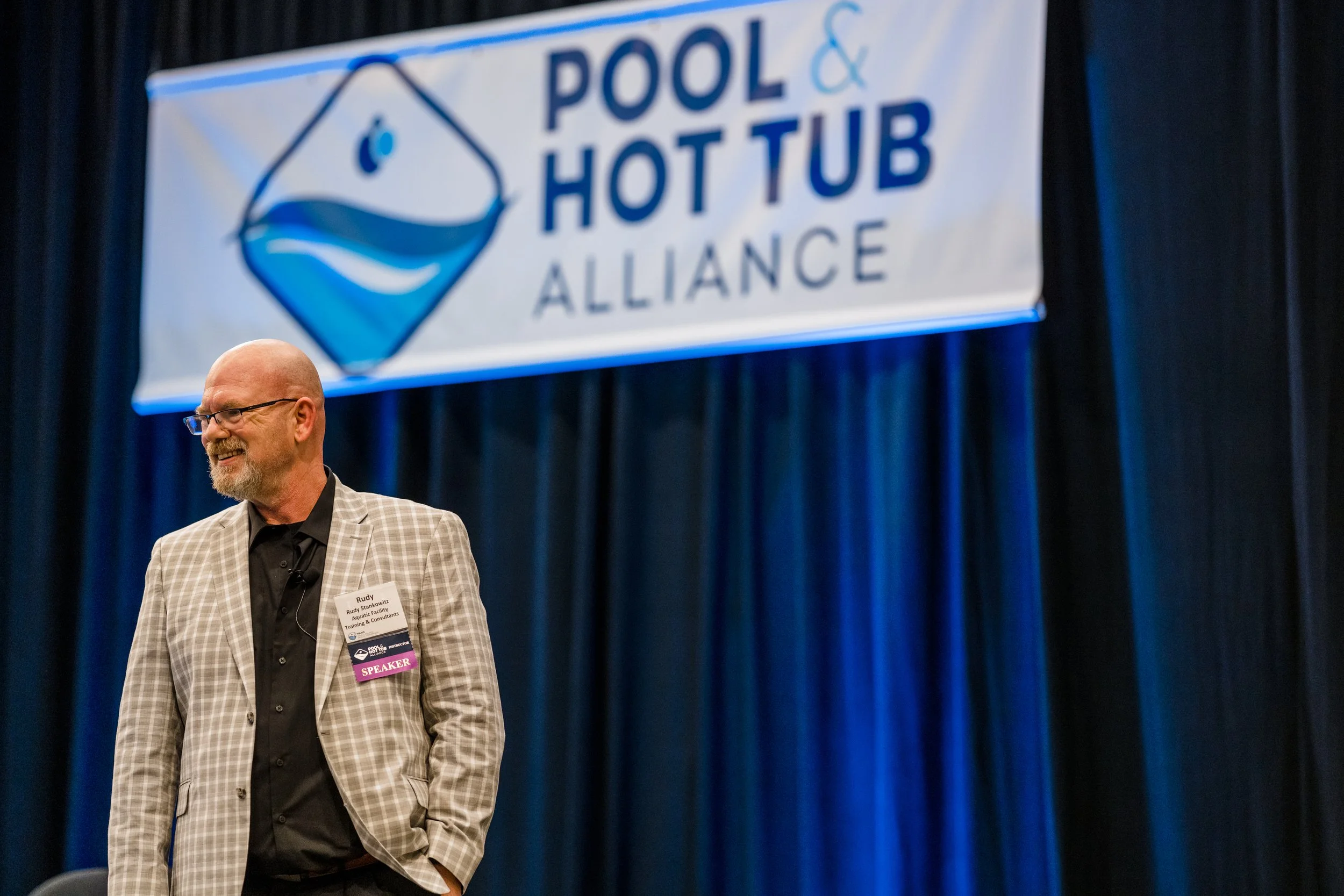 Man wearing glasses and a checkered blazer with a name tag labeled 'SPEAKER,' standing on a stage with a dark curtain background and a large banner overhead reading 'Pool & Hot Tub Alliance' with a logo of a water-filled hot tub.