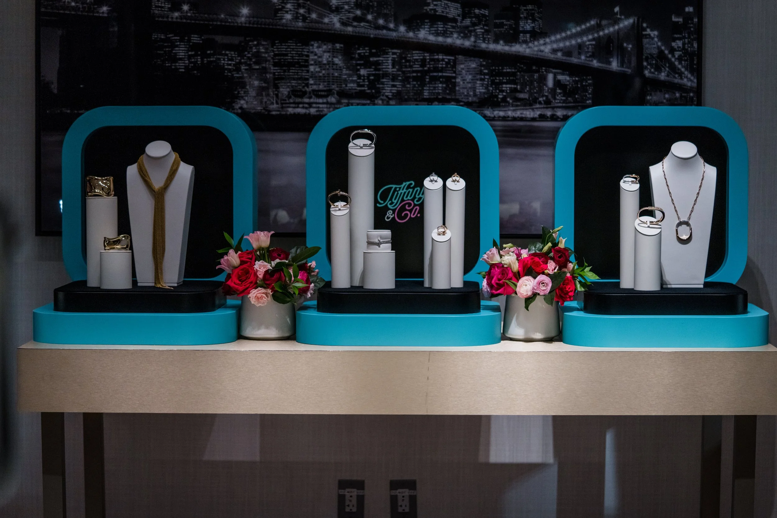 Jewelry display with rings, necklaces, and earrings showcased on white busts and cylindrical stands, adorned with pink and red flowers, with a backdrop of a cityscape at night.