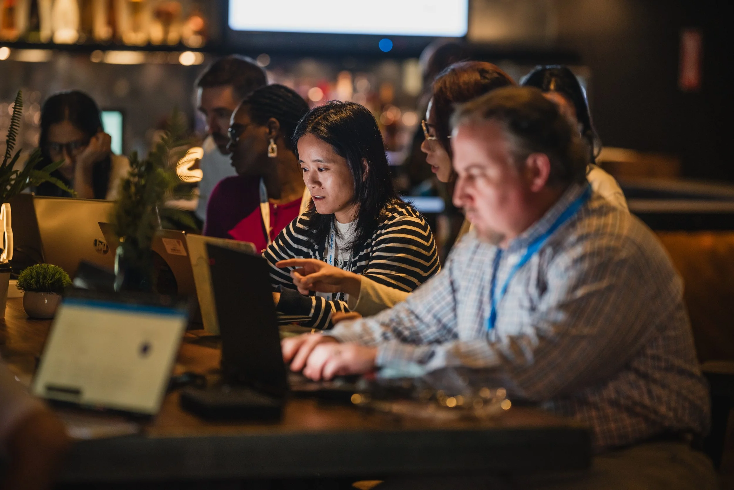 Group of diverse adults working on laptops and tablets at a crowded, dimly lit co-working space or office.