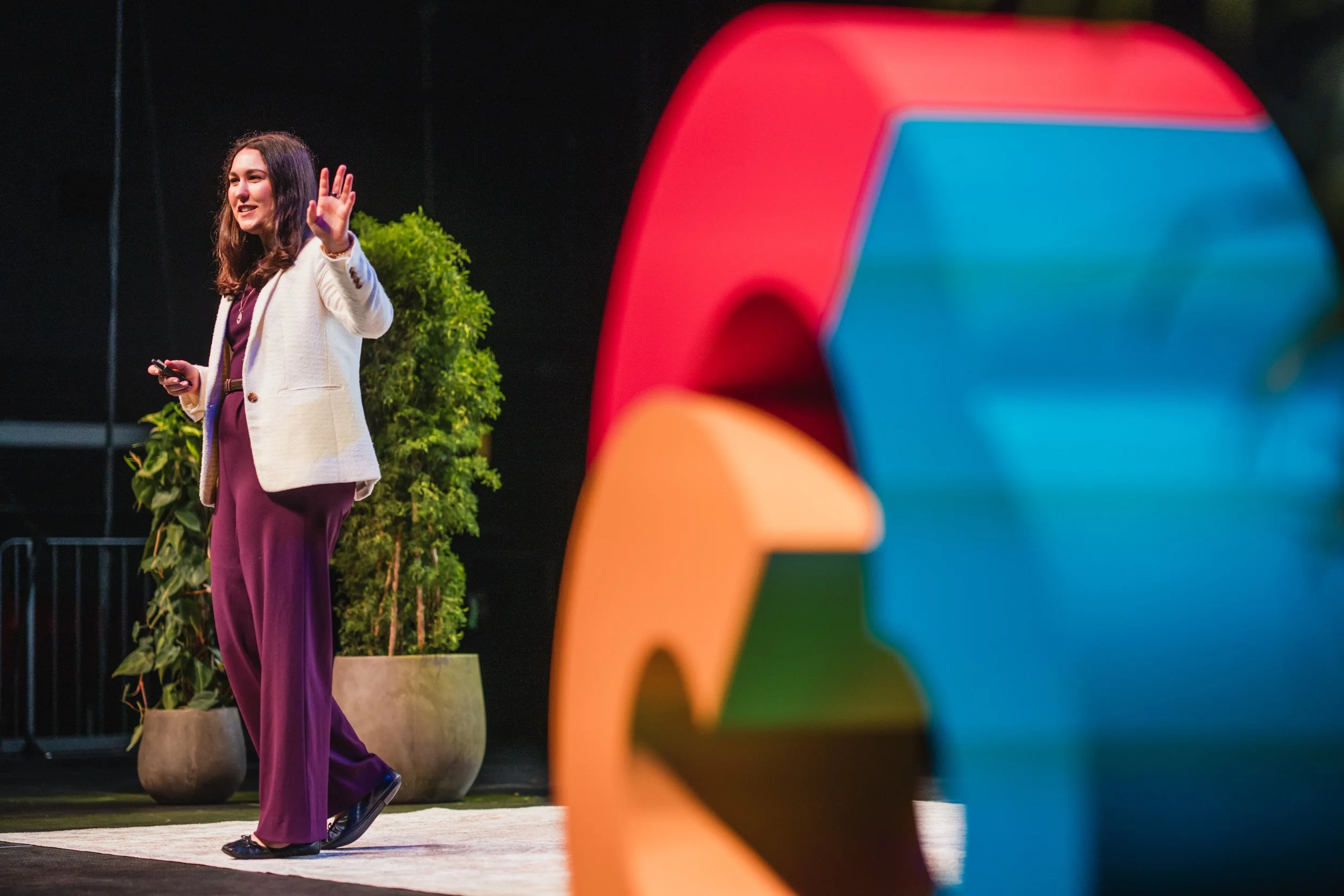 A woman in a white blazer and purple pants is giving a presentation on stage, gesturing with her hand, with colorful abstract sculptures in the foreground and greenery in the background.