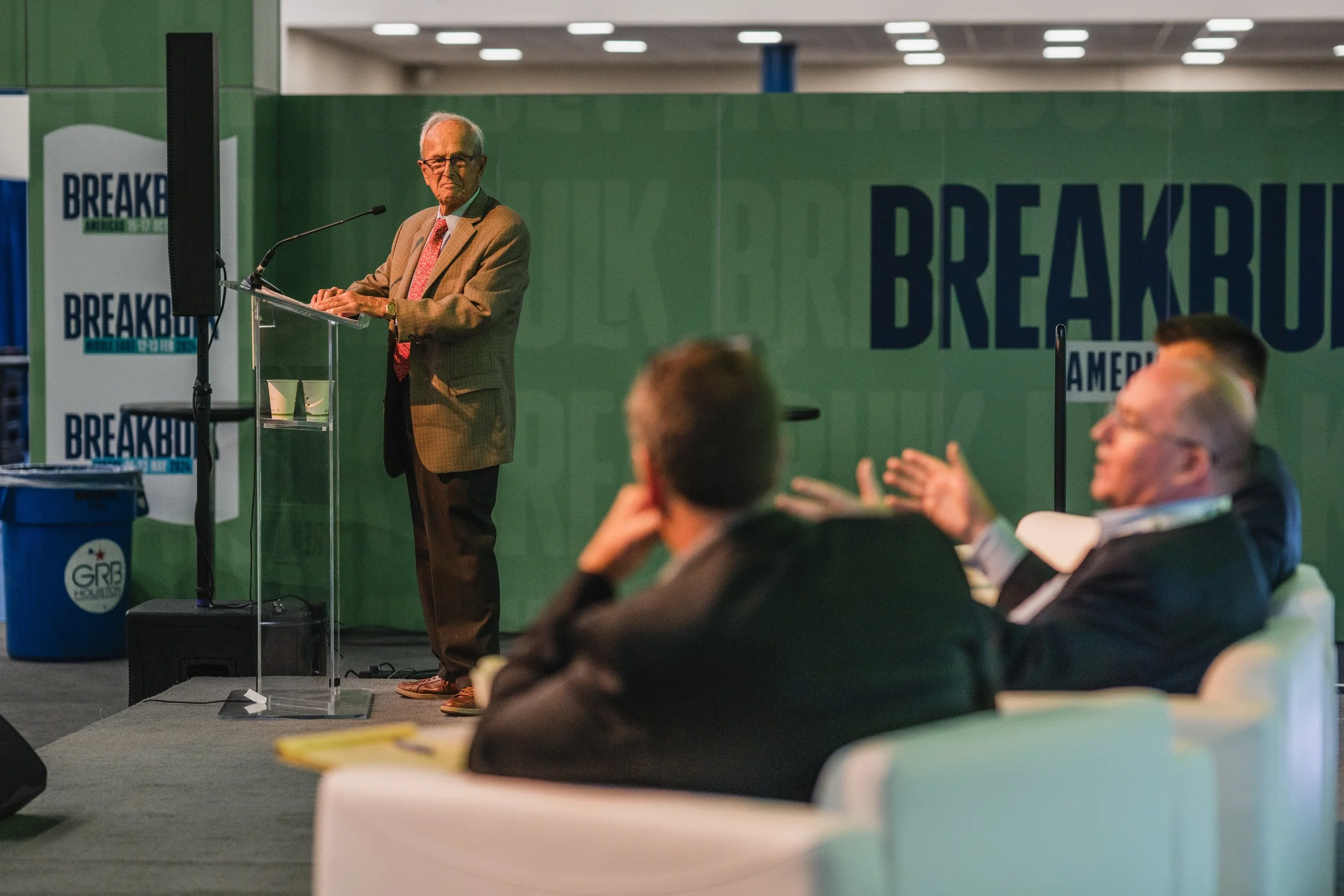 An older man in a tan blazer and red tie speaking at a podium during a political event, with a green background displaying the word 'BREAKB' and seated audience members clapping.