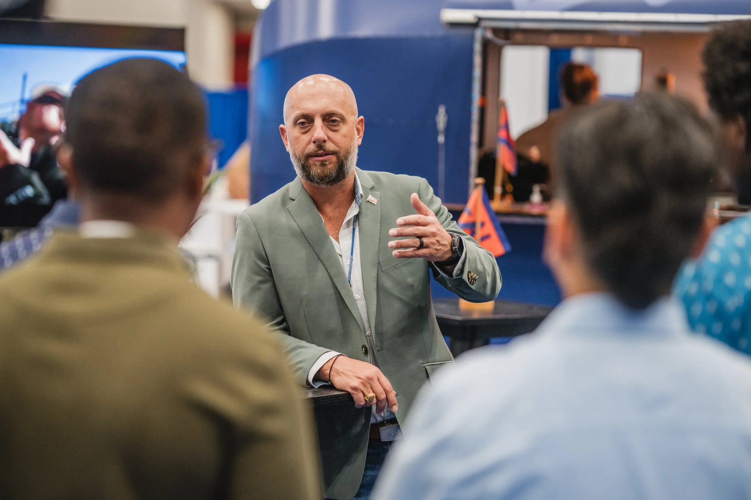 A man with a bald head and gray beard wearing a light green blazer and a watch, speaking to a group of people indoors, with some American flags in the background.