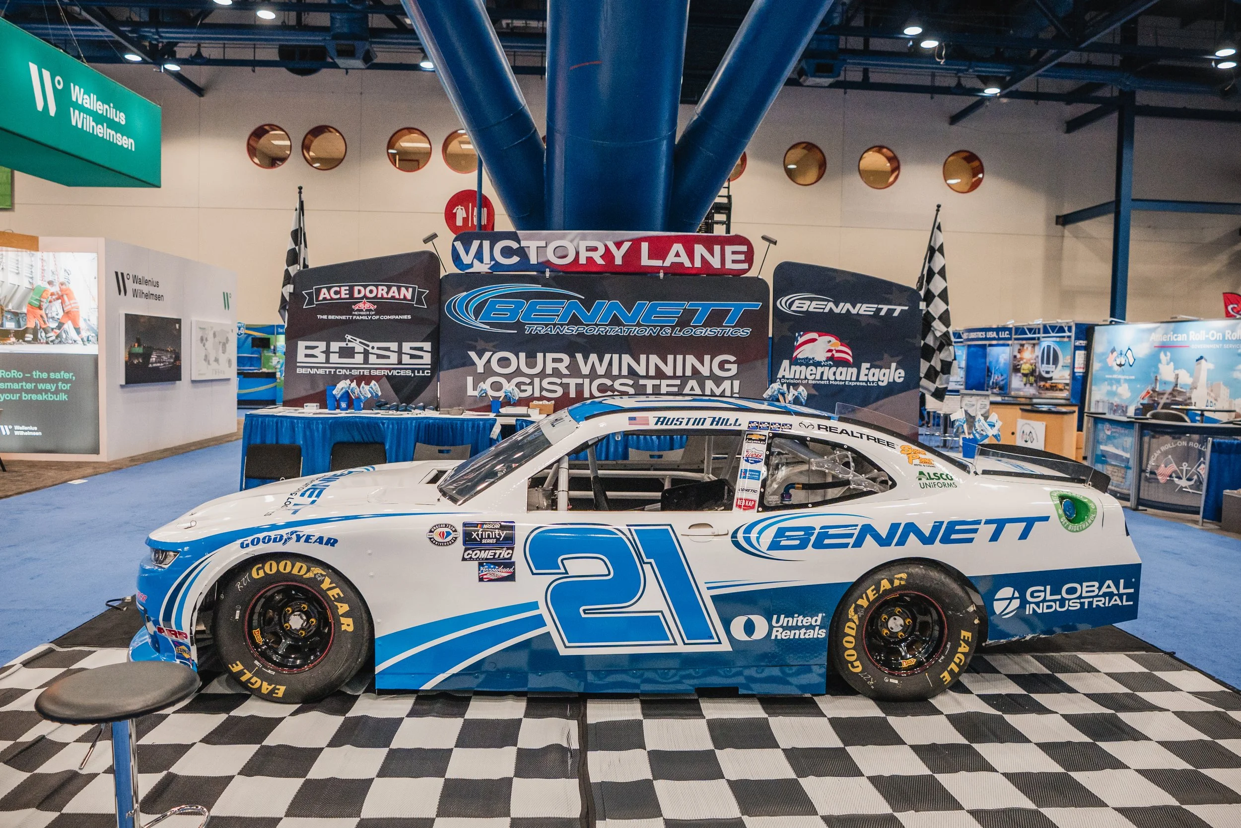 Race car with the number 21, branded with Bennet and other sponsors, displayed in an exhibition hall with banners and signs promoting logistics companies. The car is positioned on a checkered platform in front of a blue structure, with a seating area