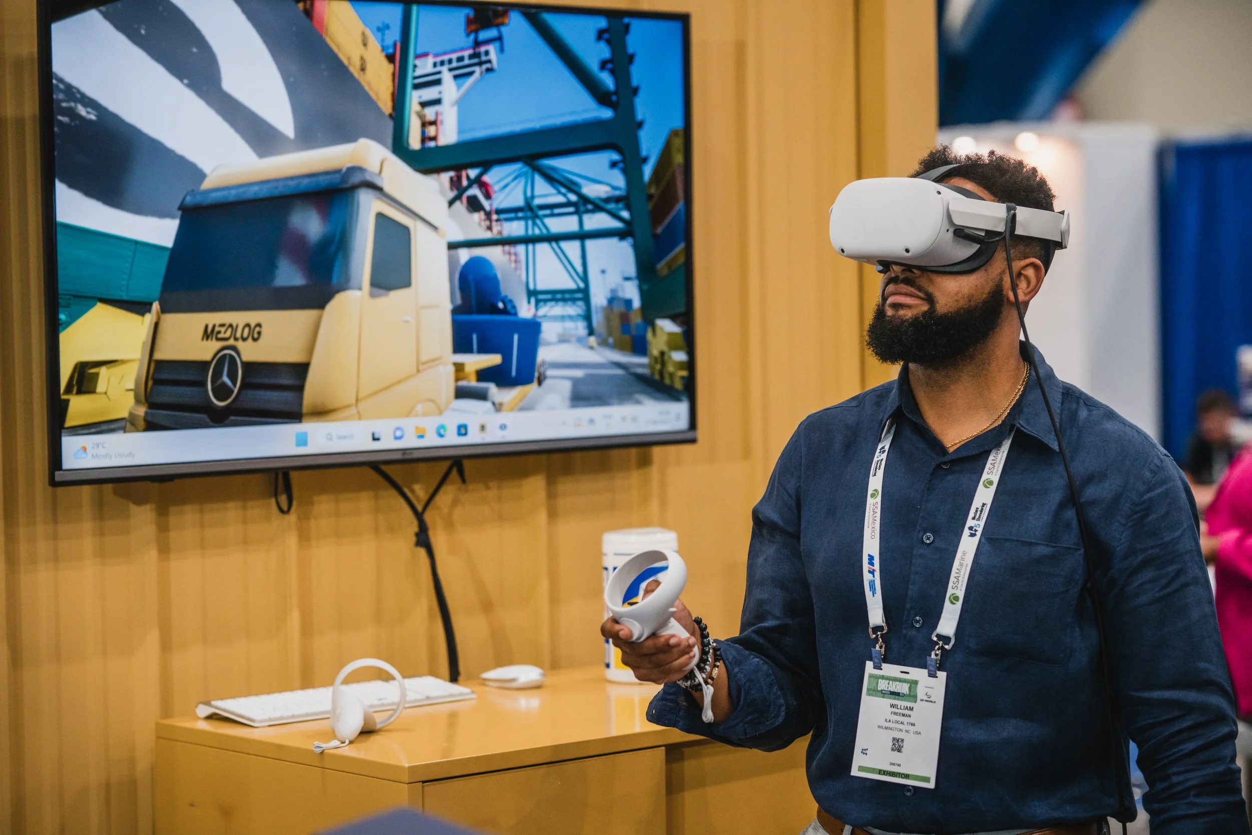 A man with a beard using a virtual reality headset and controller at a booth, with a display screen showing a cartoon or animated scene of a yellow truck labeled 'MEDLOG' in an industrial or warehouse setting.
