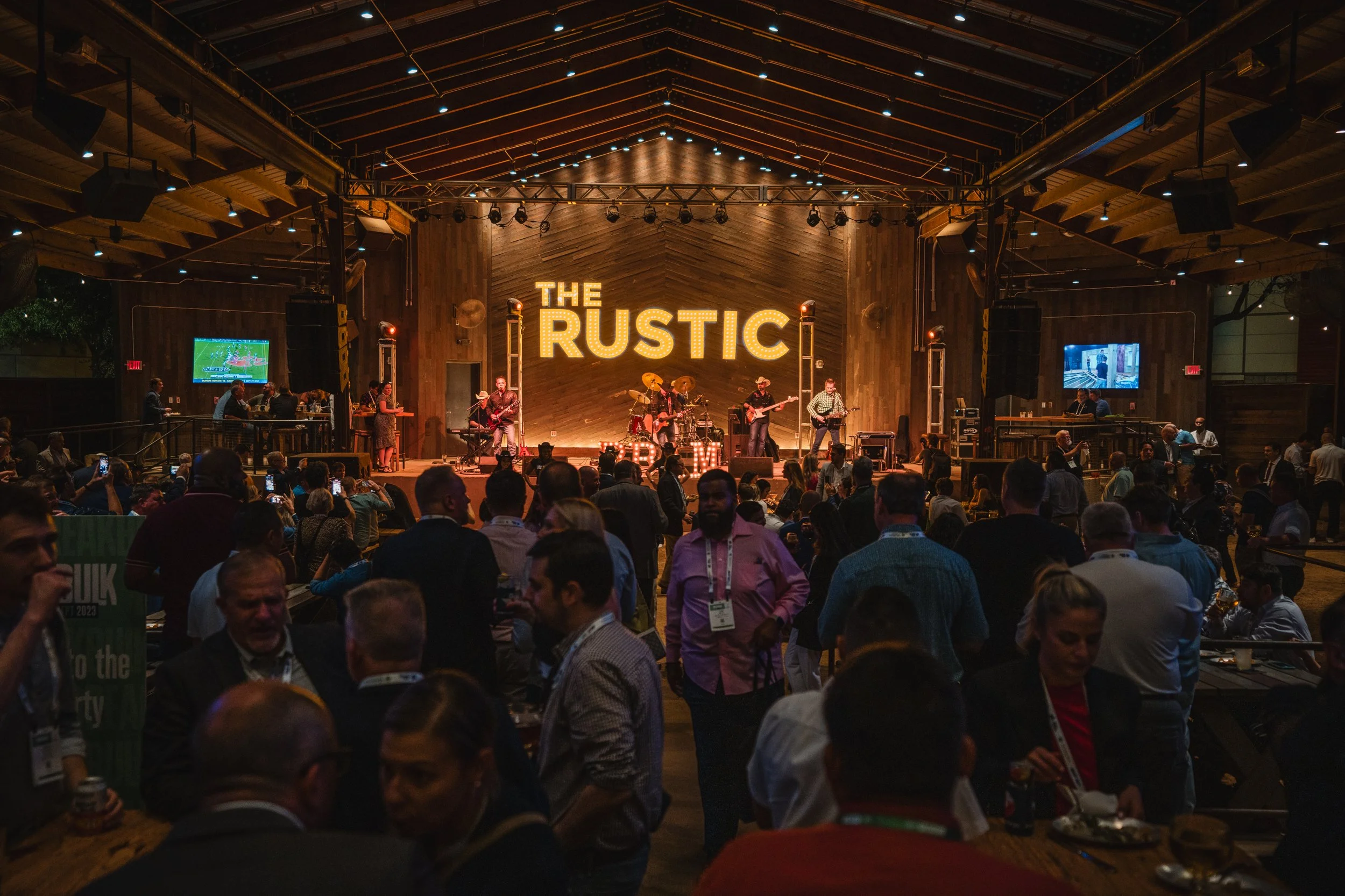 Live music performance on stage at an event called 'The Rustic' with an audience standing and socializing in a warm, rustic indoor venue.