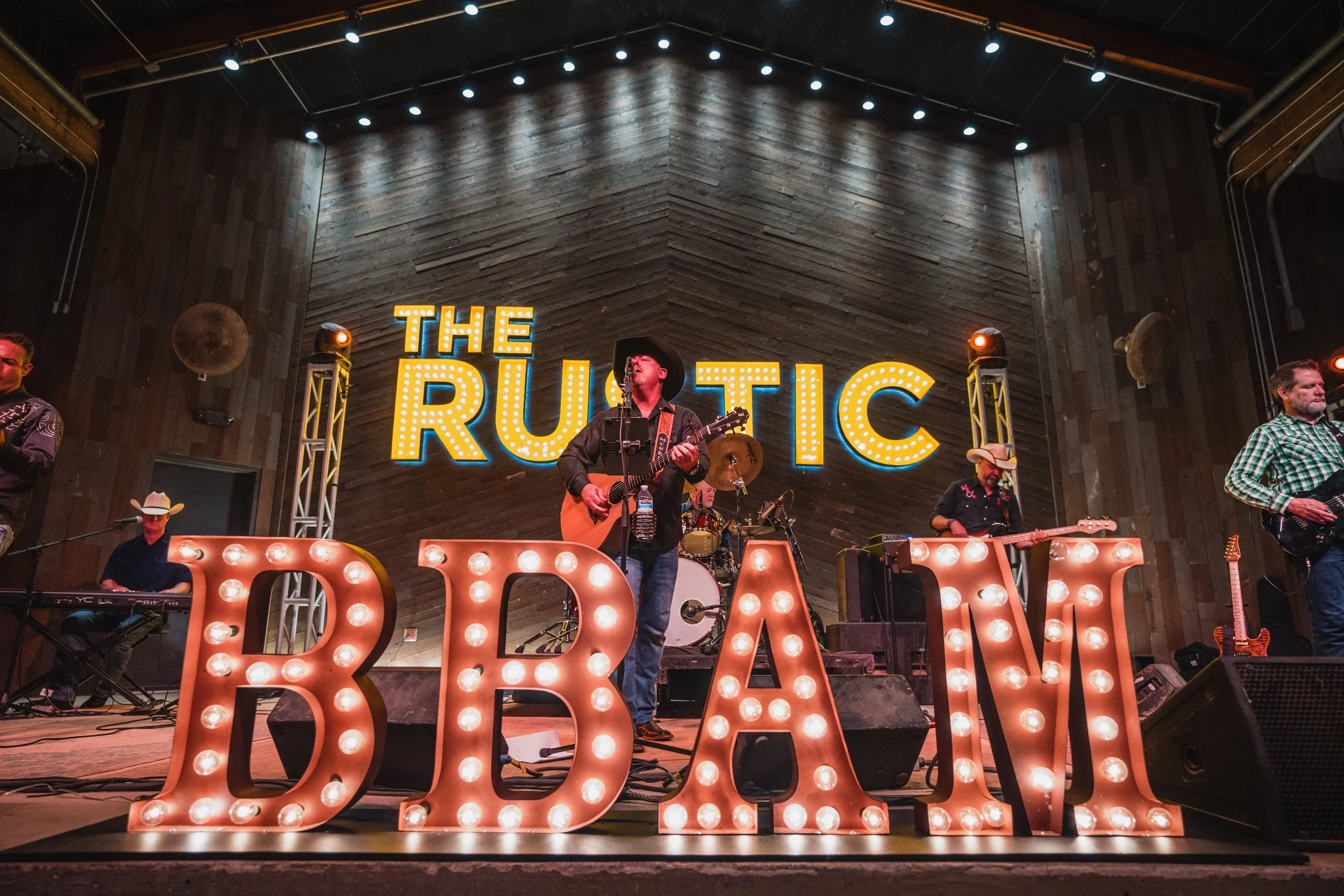 A band performing on stage under a sign that reads 'The Rustic'. The stage has large illuminated letters spelling 'BARM' in the foreground, and the musicians include a guitarist, a keyboard player wearing a cowboy hat, and a drummer.