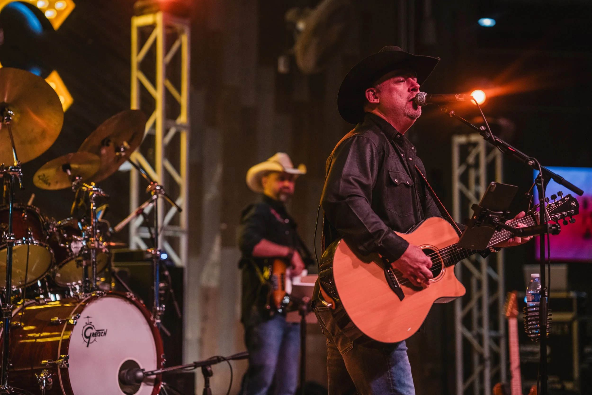 A country band performing on stage, with a man in a cowboy hat playing an acoustic guitar and singing into a microphone, another man in a cowboy hat playing bass guitar in the background, and drums to the left, under stage lights.
