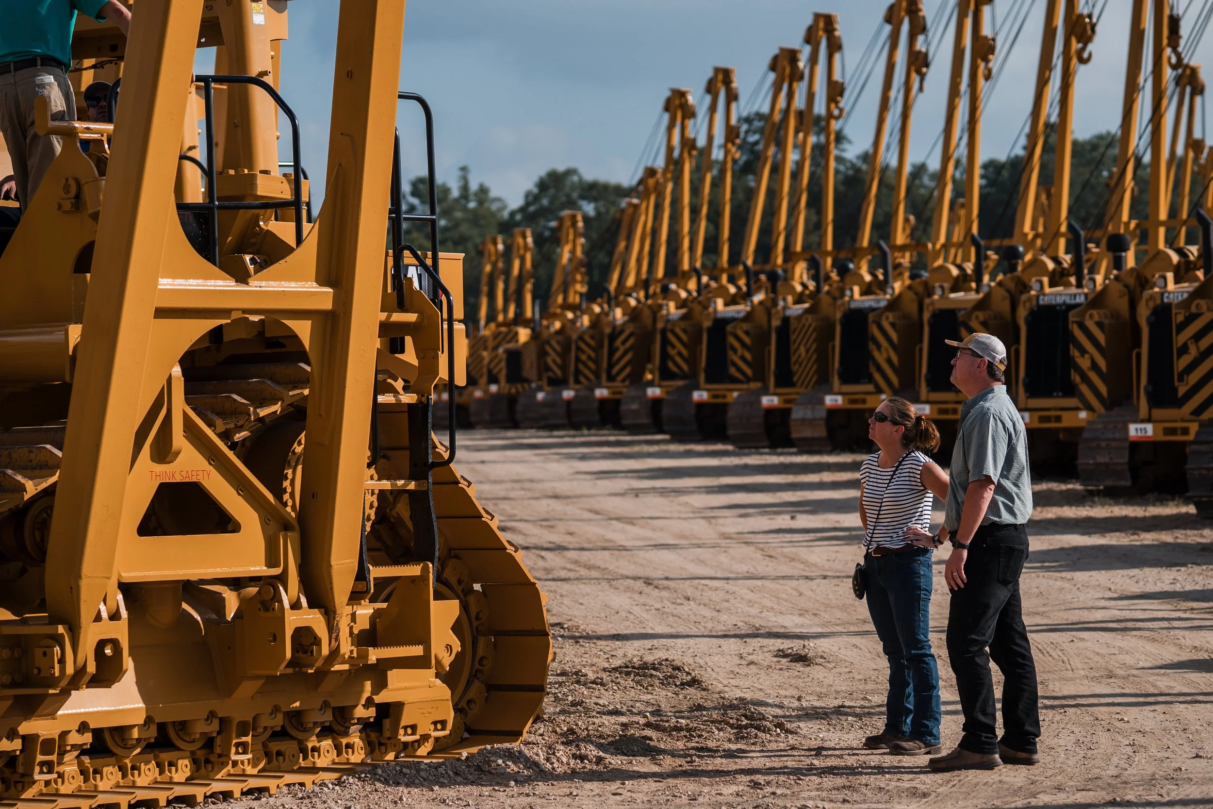 A man and woman looking at yellow bulldozers lined up on a dirt area.