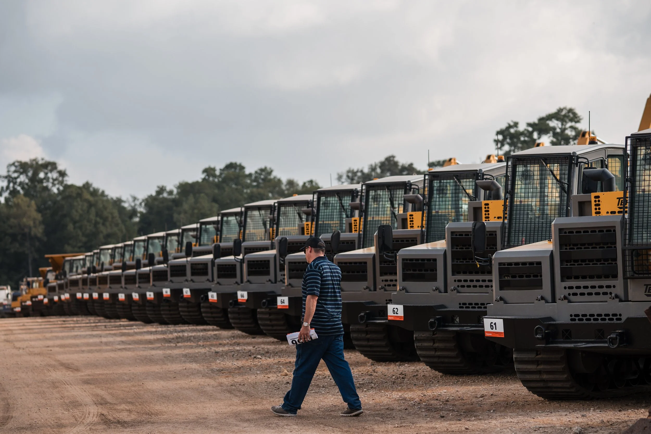 A man walking past a row of construction vehicles parked on a dirt lot, with trees in the background and an overcast sky.