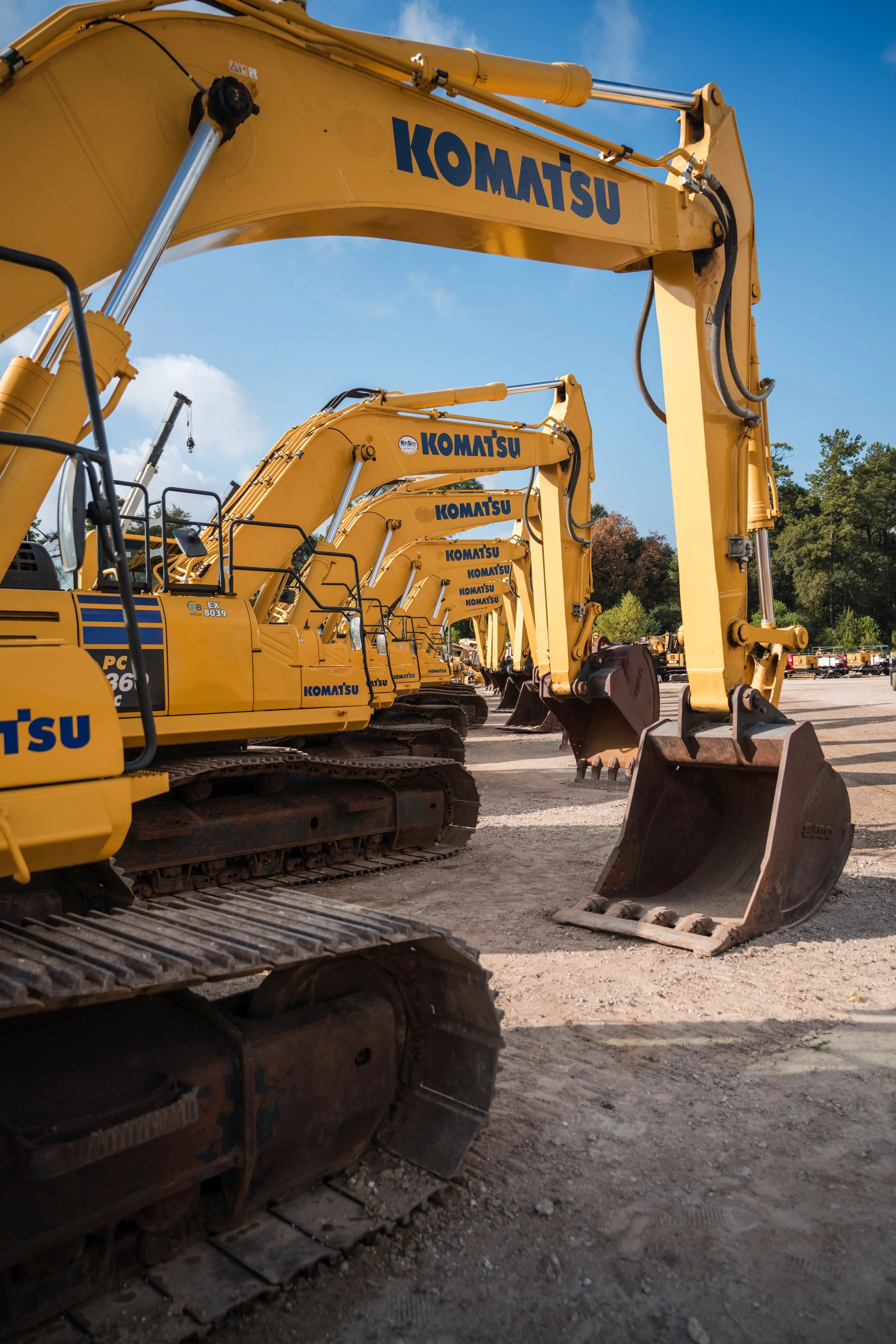 A row of yellow Komatsu excavators parked on a construction site, with blue skies and trees in the background.
