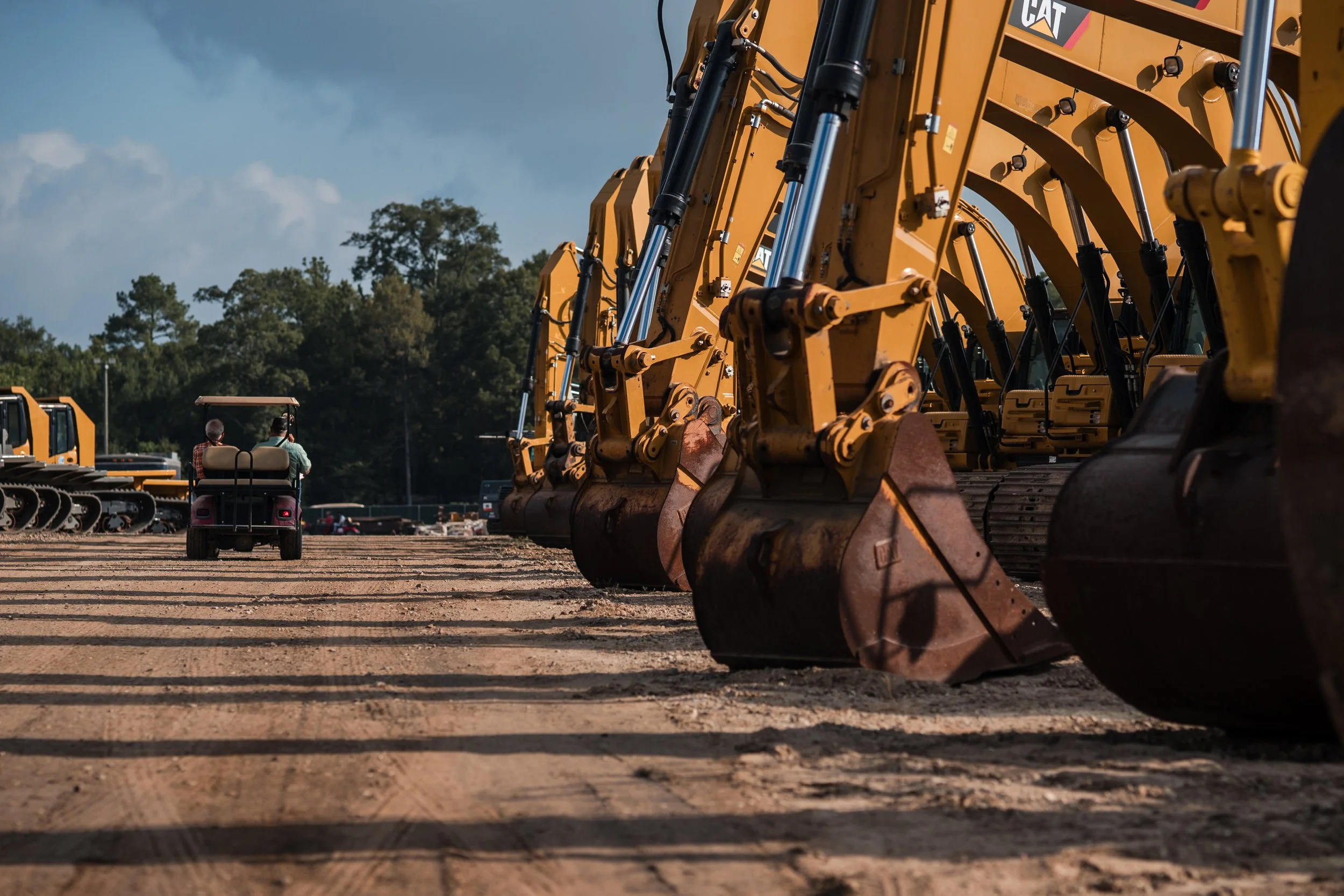 Row of yellow construction excavators on a dirt lot with two people riding in a golf cart in the background.