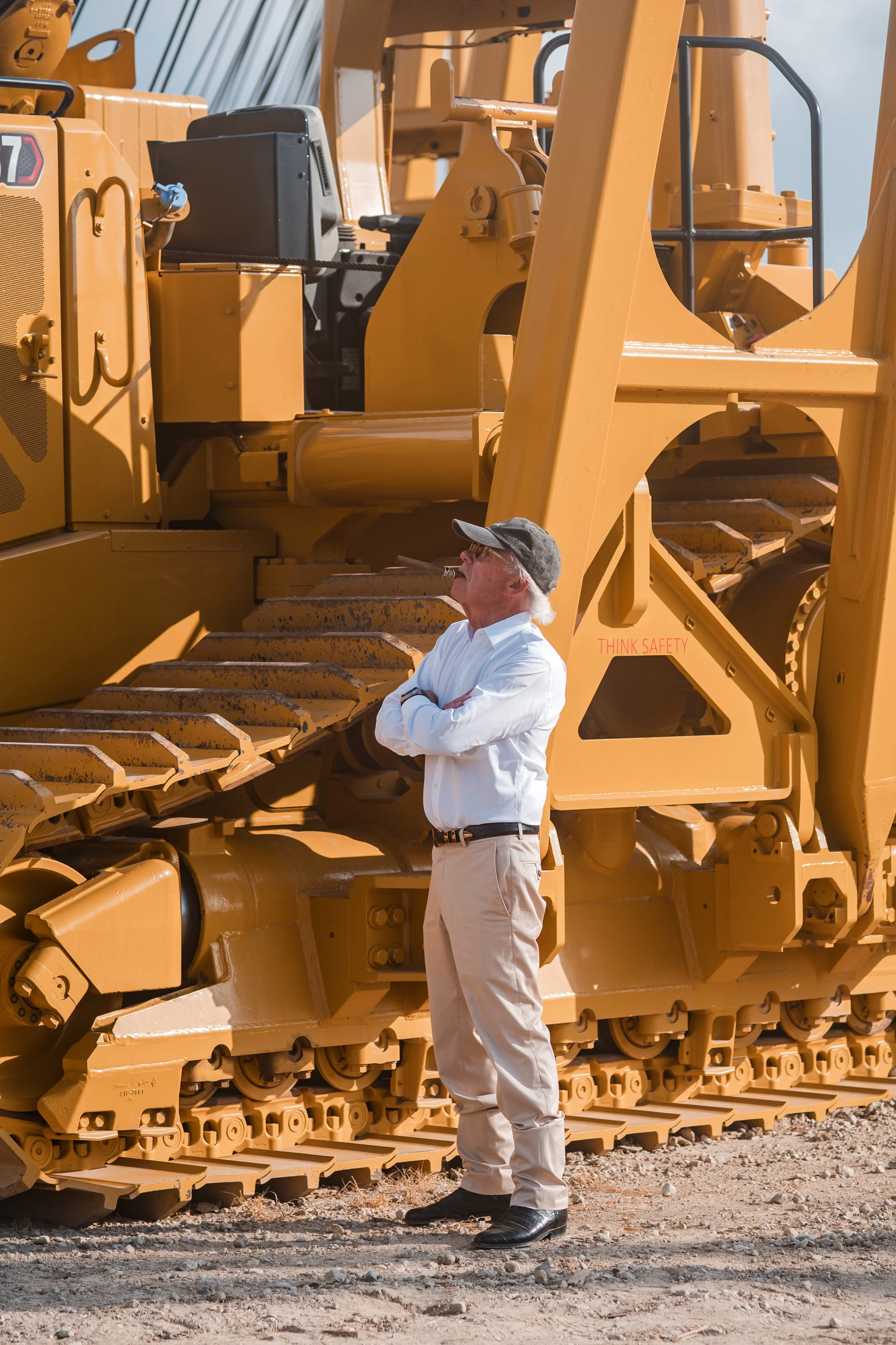 A man with white hair and a gray cap standing with arms crossed next to a large yellow construction bulldozer. The man is wearing a white shirt, beige pants, and black shoes, and appears to be inspecting or contemplating the machinery.