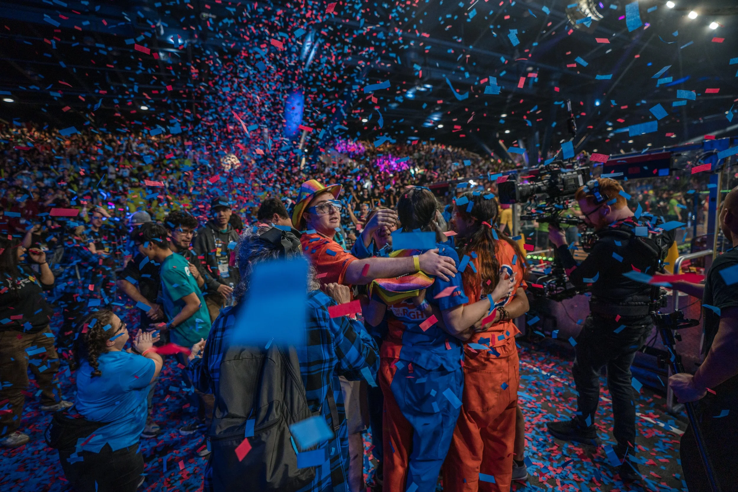 Celebration with confetti falling at a large indoor event, with a crowd of people hugging and a camera crew recording.