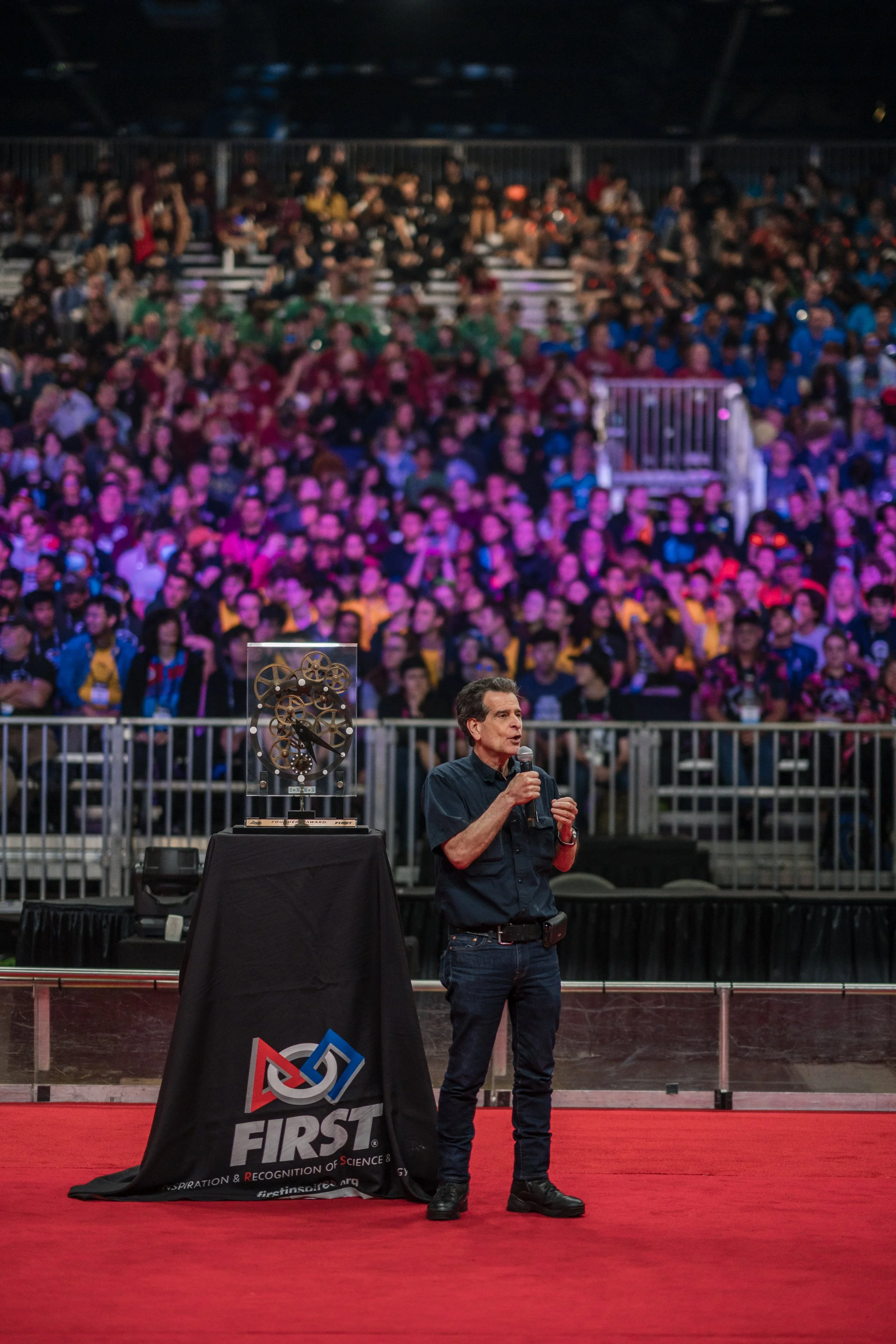 A man holding a microphone stands next to a display case with mechanical gears at a FIRST robotics event, with a large seated audience in the background.