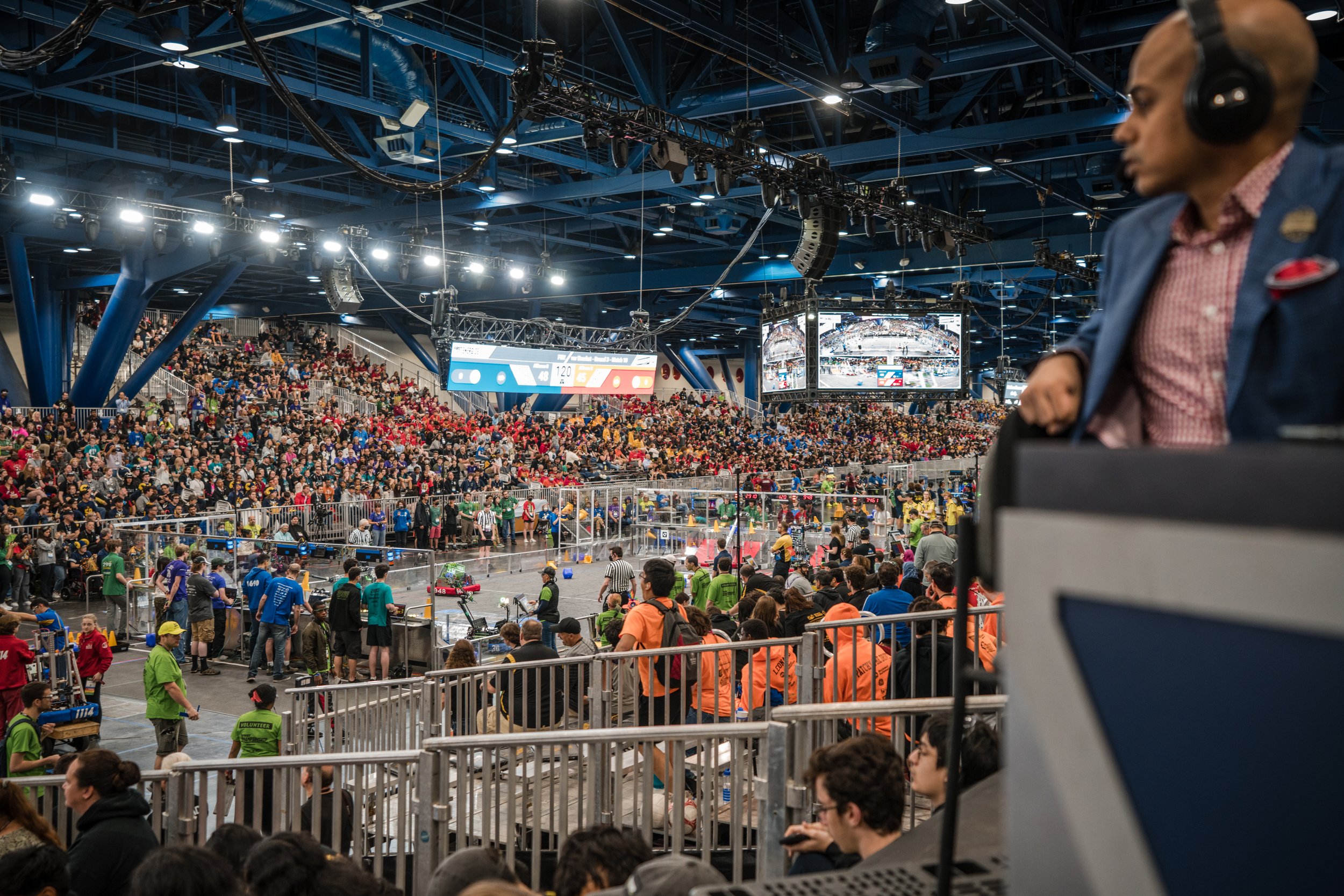 Indoor robotics competition with a crowd of spectators watching the event, volunteers, and a judge at a table in the foreground.