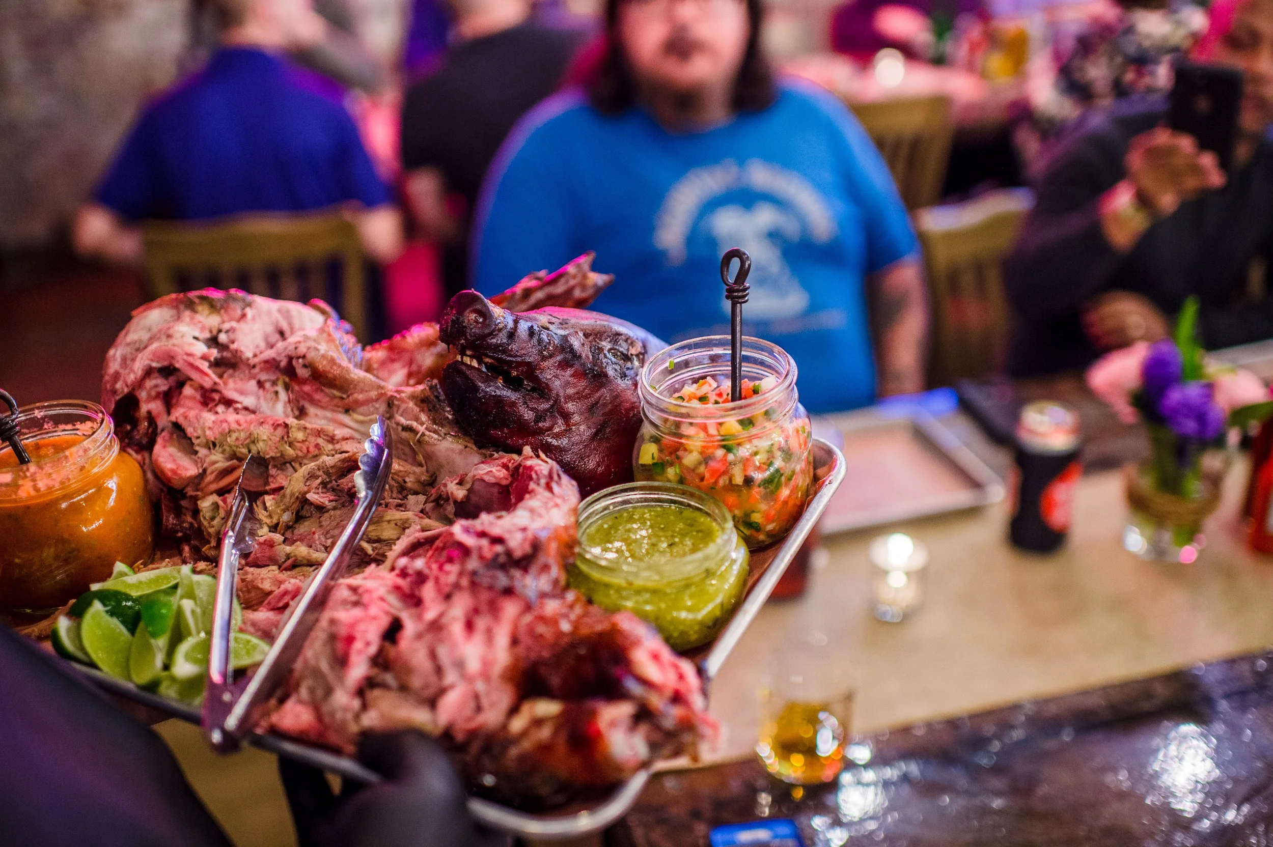 A serving tray with different cuts of cooked meat, including a whole animal's head, accompanied by lime wedges, various sauces, and chopped vegetables at a restaurant or dinner event.