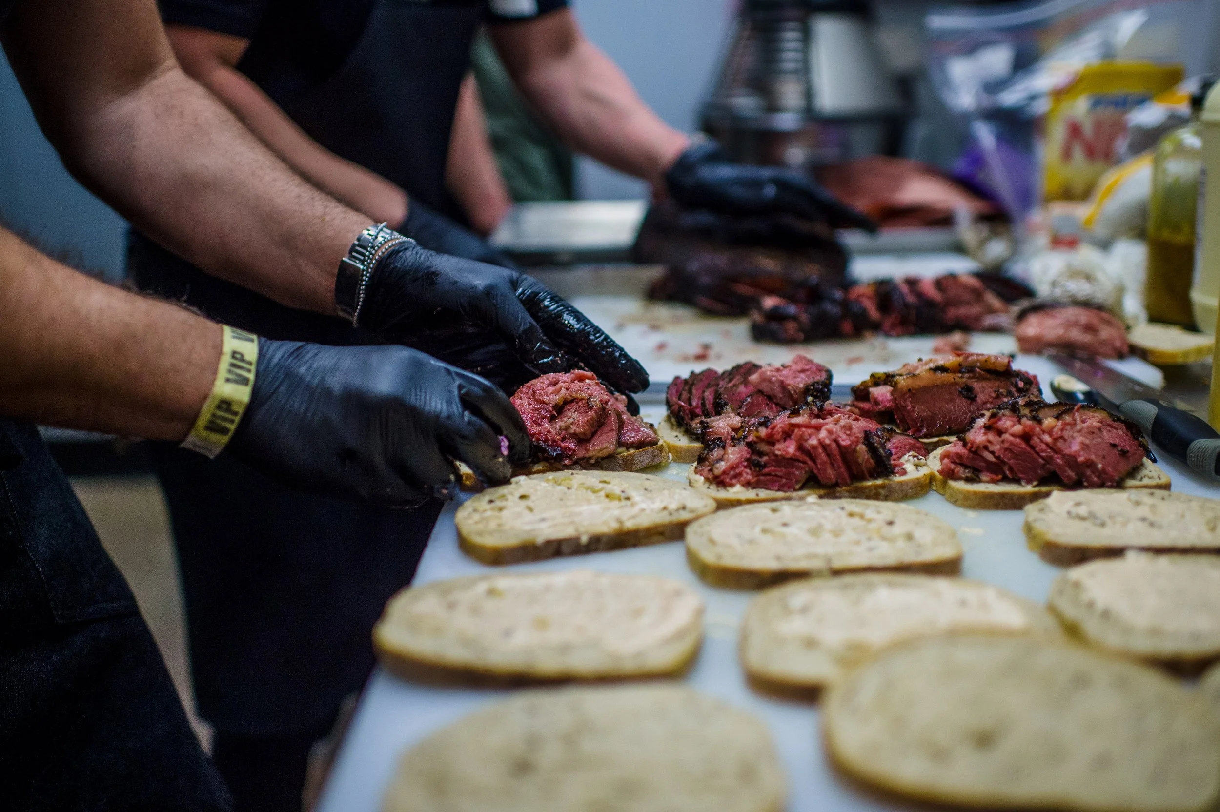 People preparing sandwiches with sliced bread and sliced beef on a table, wearing black gloves.