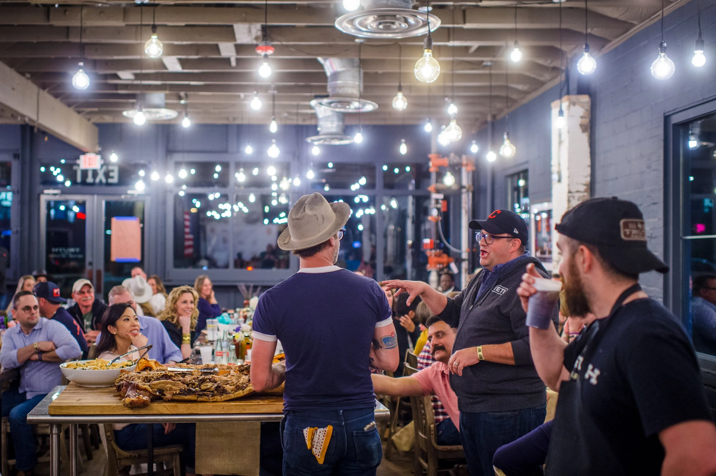 A group of people gathered in a restaurant, with three men standing and talking near a table with food, while others are seated and listening. The restaurant has a casual atmosphere with exposed ceiling beams and hanging light bulbs.