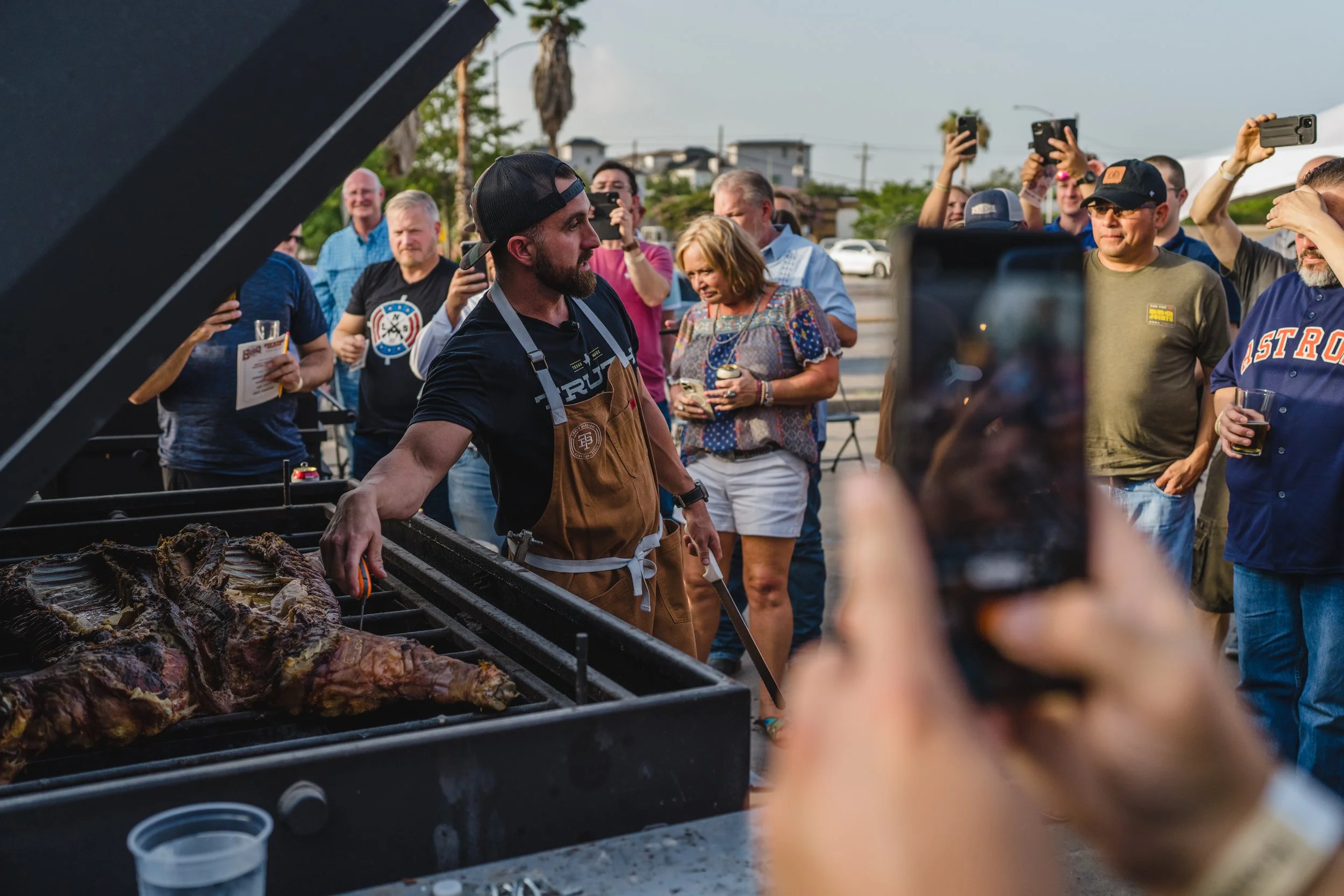 A man wearing a brown apron grills large pieces of meat at an outdoor event, with a crowd of people around him taking photos and holding drinks.