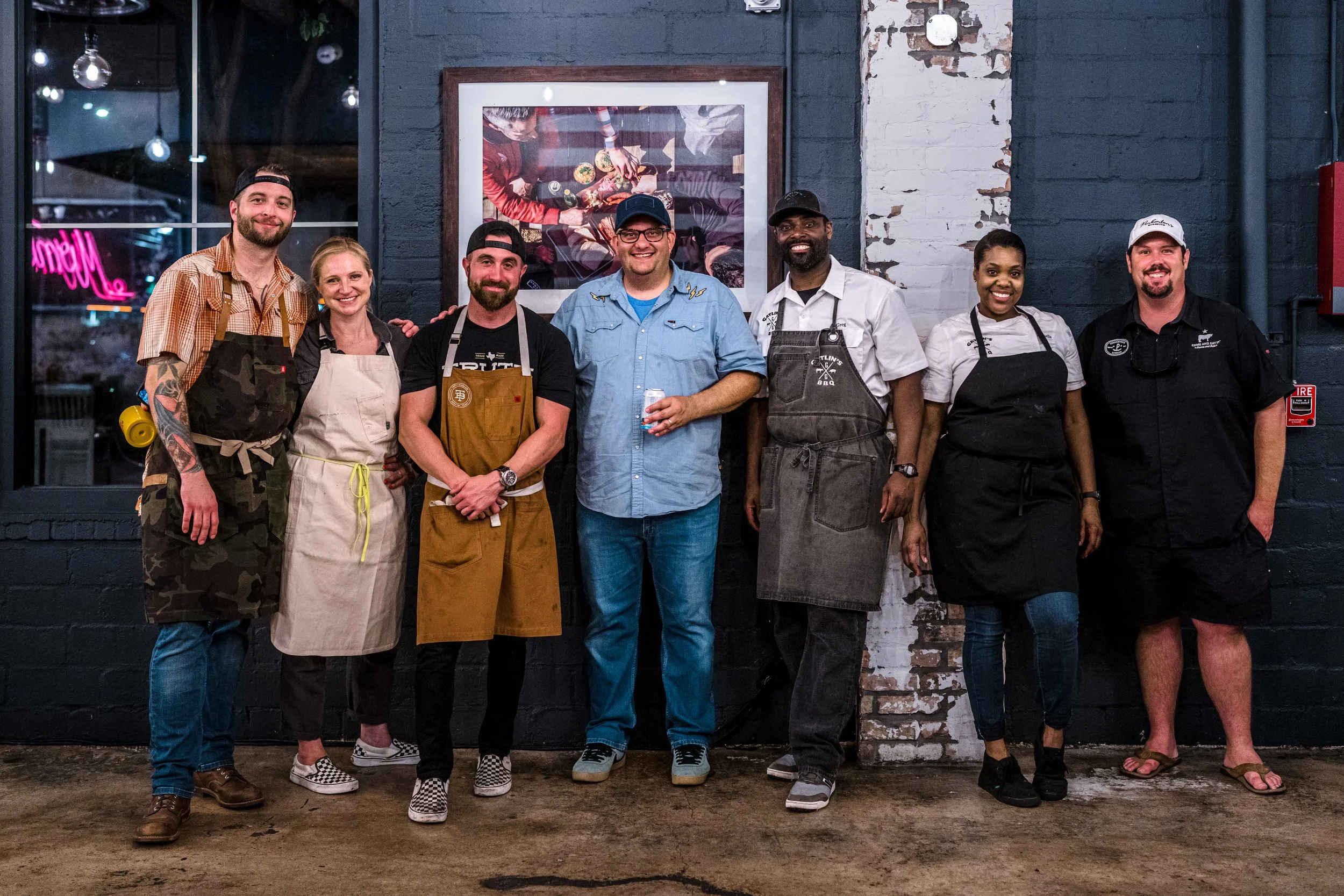Group of seven diverse restaurant staff members standing side by side inside a restaurant, dressed in aprons and casual clothing, smiling at the camera.