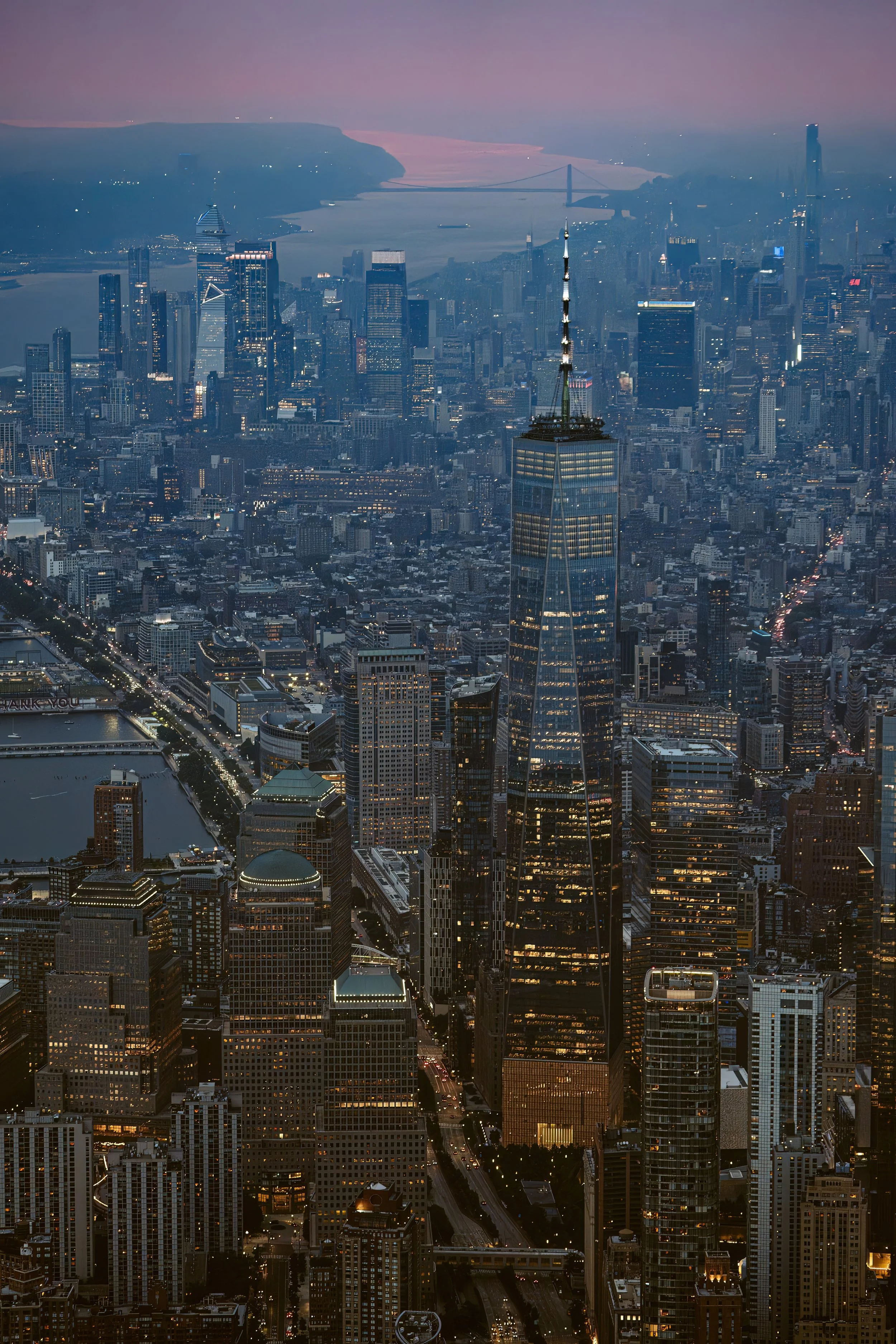 Widow View of New York City skyline at dusk, featuring One World Trade Center and a view of the Brooklyn Bridge in the background.