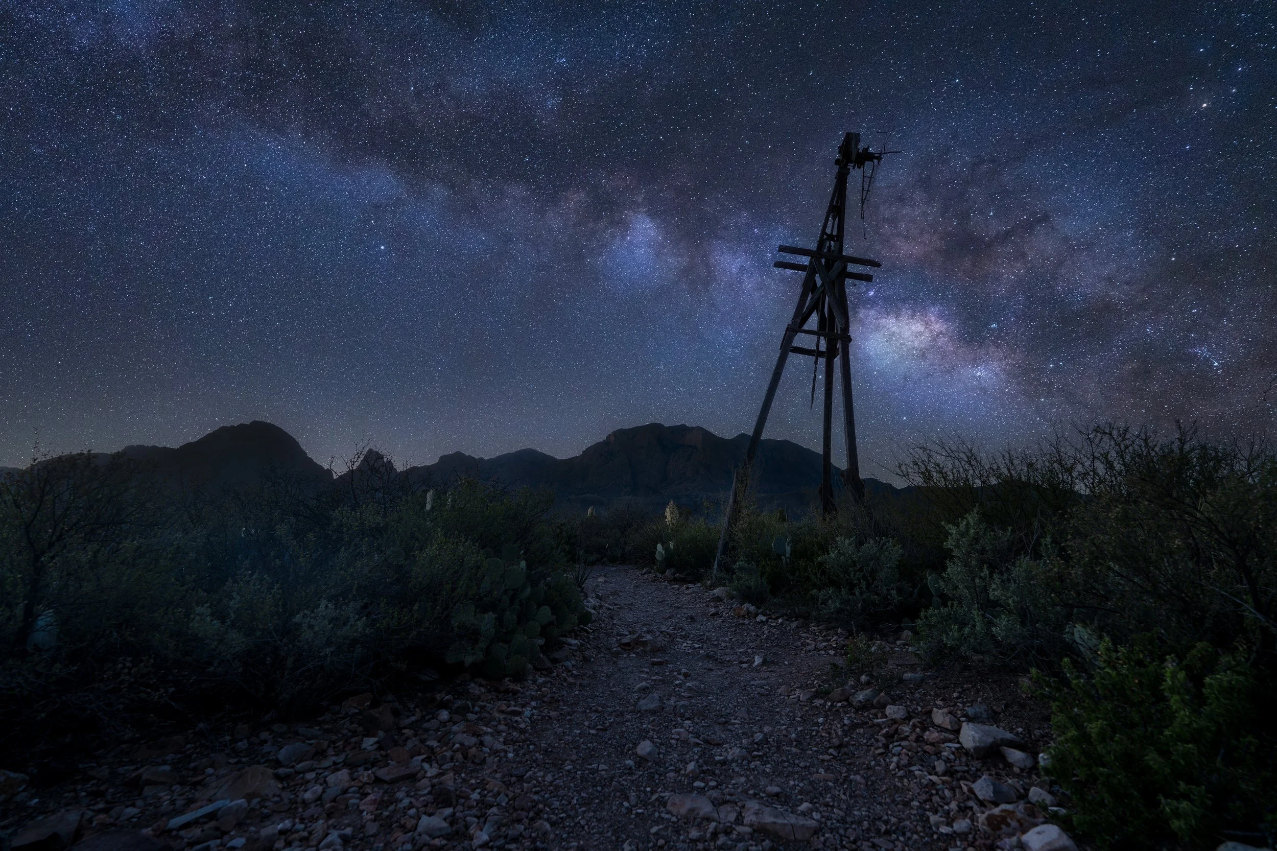 Nighttime desert landscape with a dirt trail, cacti, bushes, mountains in the background, a wooden power pole, and a star-filled sky with the Milky Way galaxy.