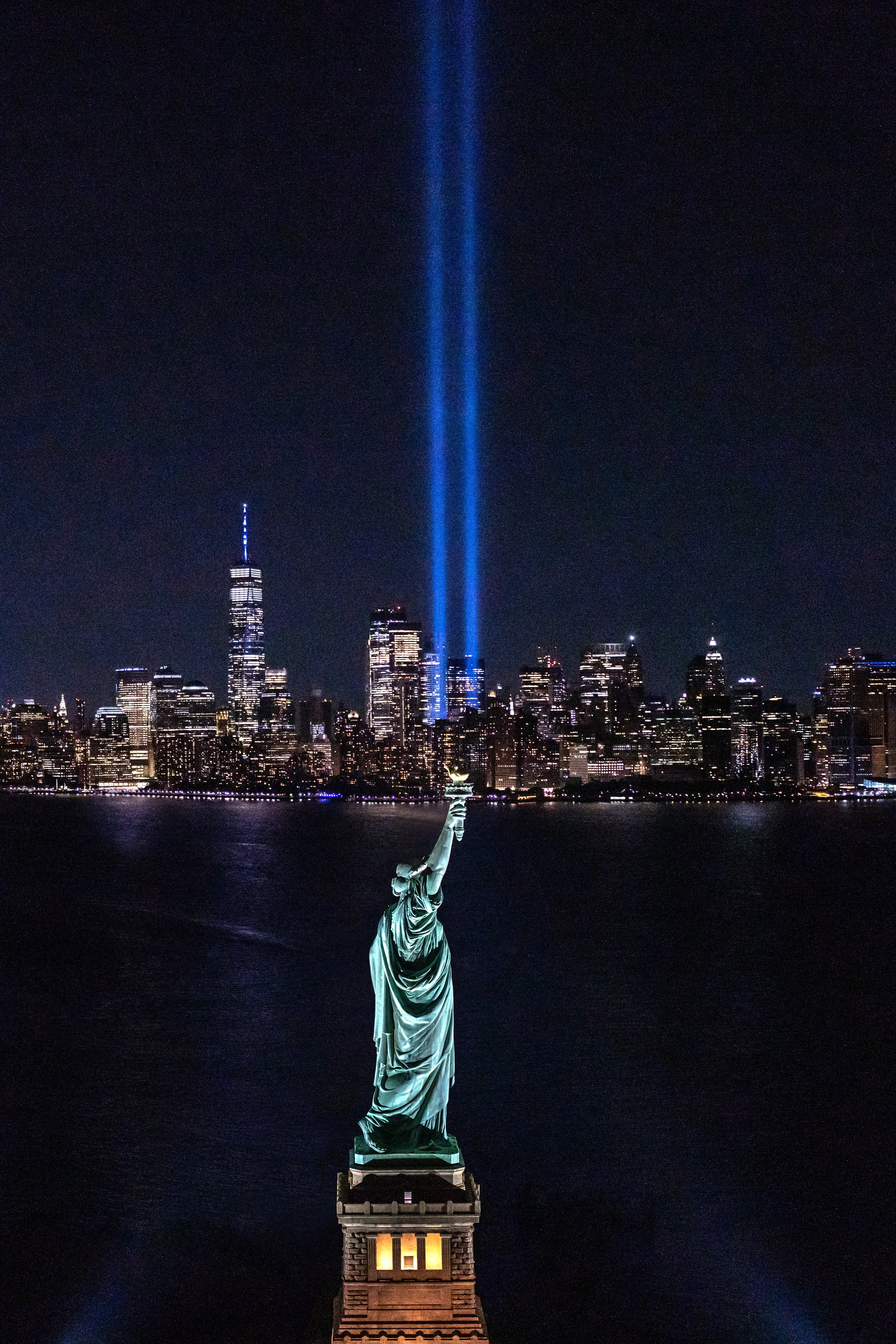 Nighttime view of the Statue of Liberty with blue tribute lights in the sky and the New York City skyline in the background.