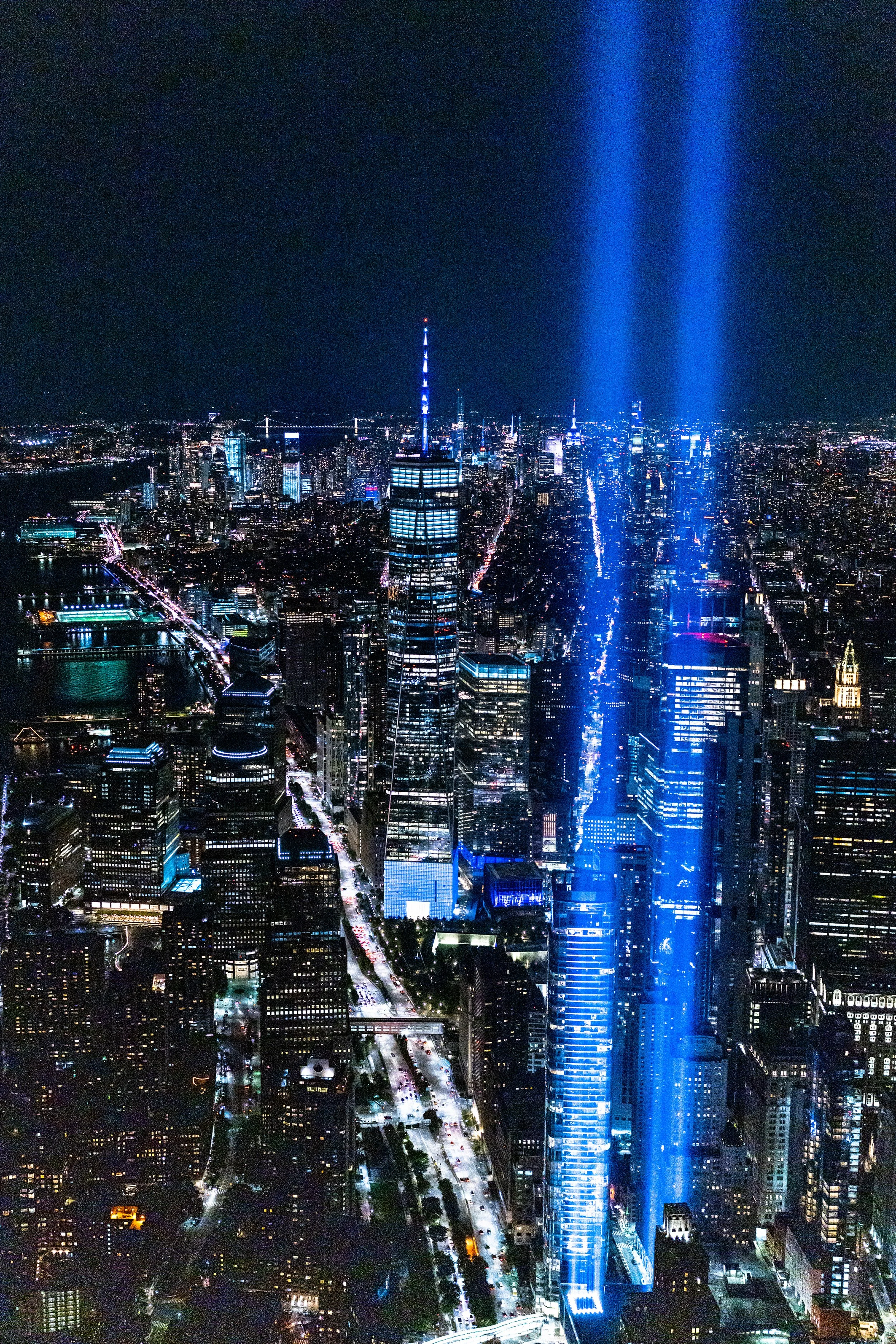 Nighttime aerial view of New York City skyline with illuminated buildings and blue light beams projecting into the sky.