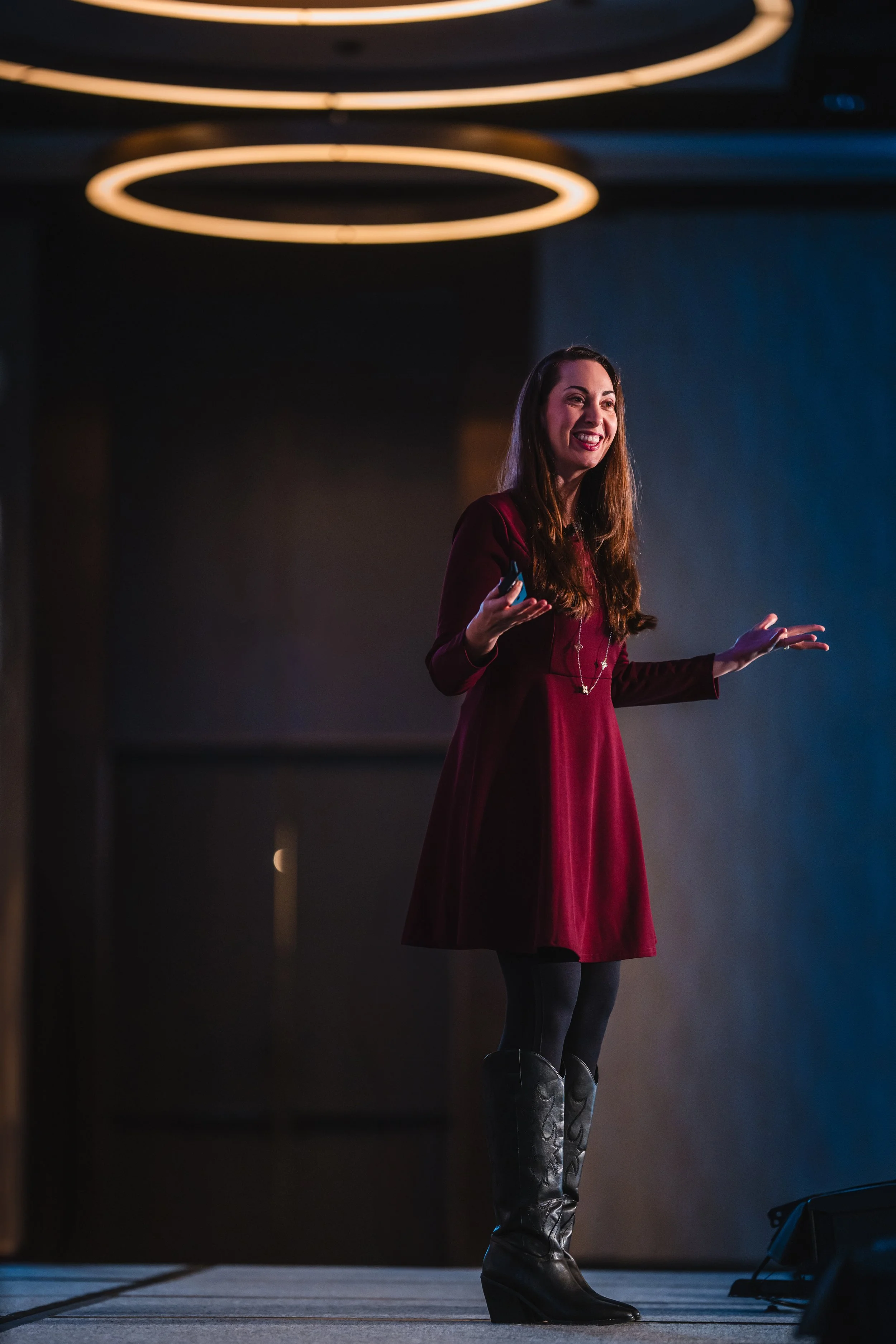A woman giving a presentation on a stage, wearing a red dress and black knee-high boots, with circular ceiling lights above her.