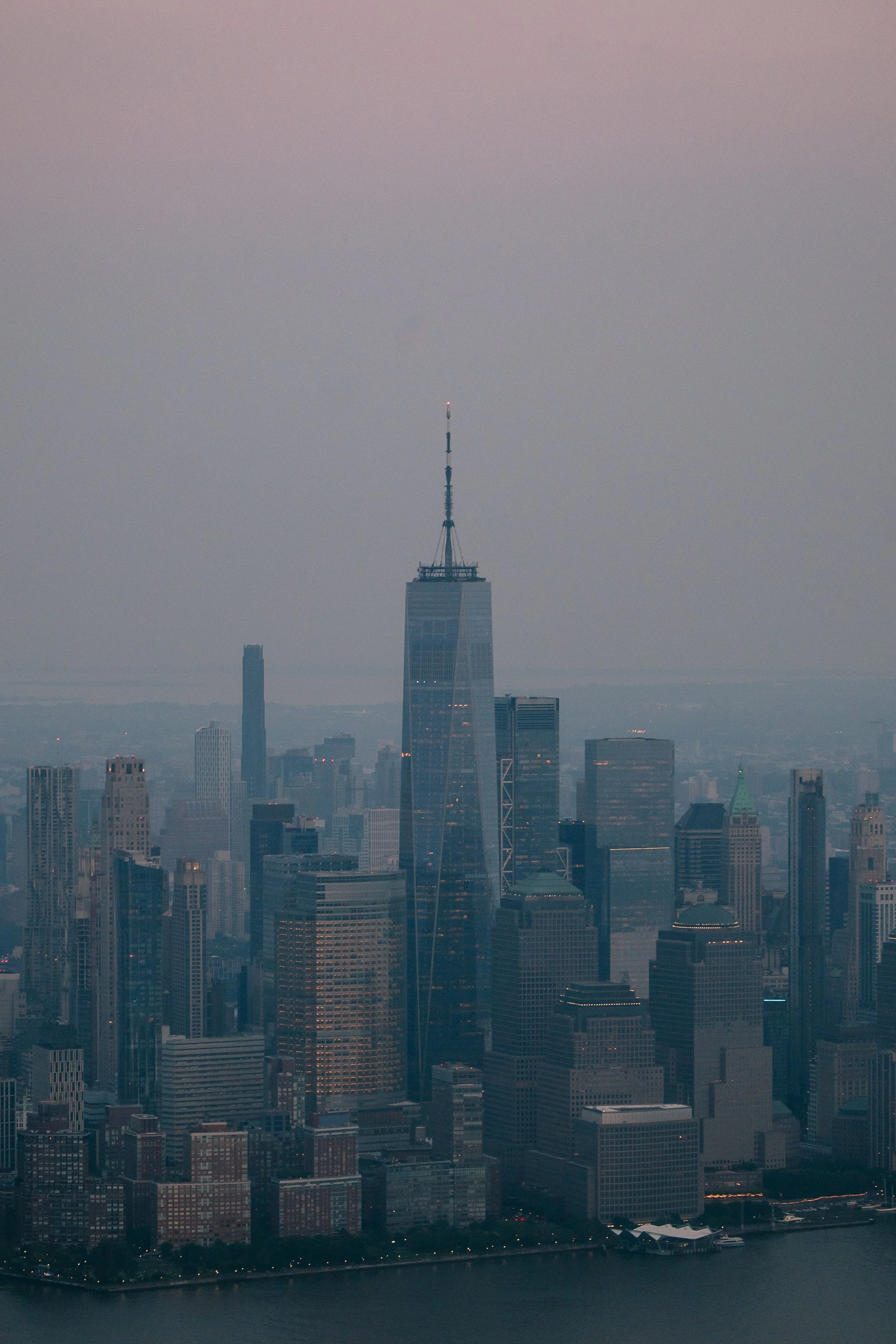 View of Manhattan skyline featuring the One World Trade Center in New York City at dusk, with a cloudy sky and water in the foreground.