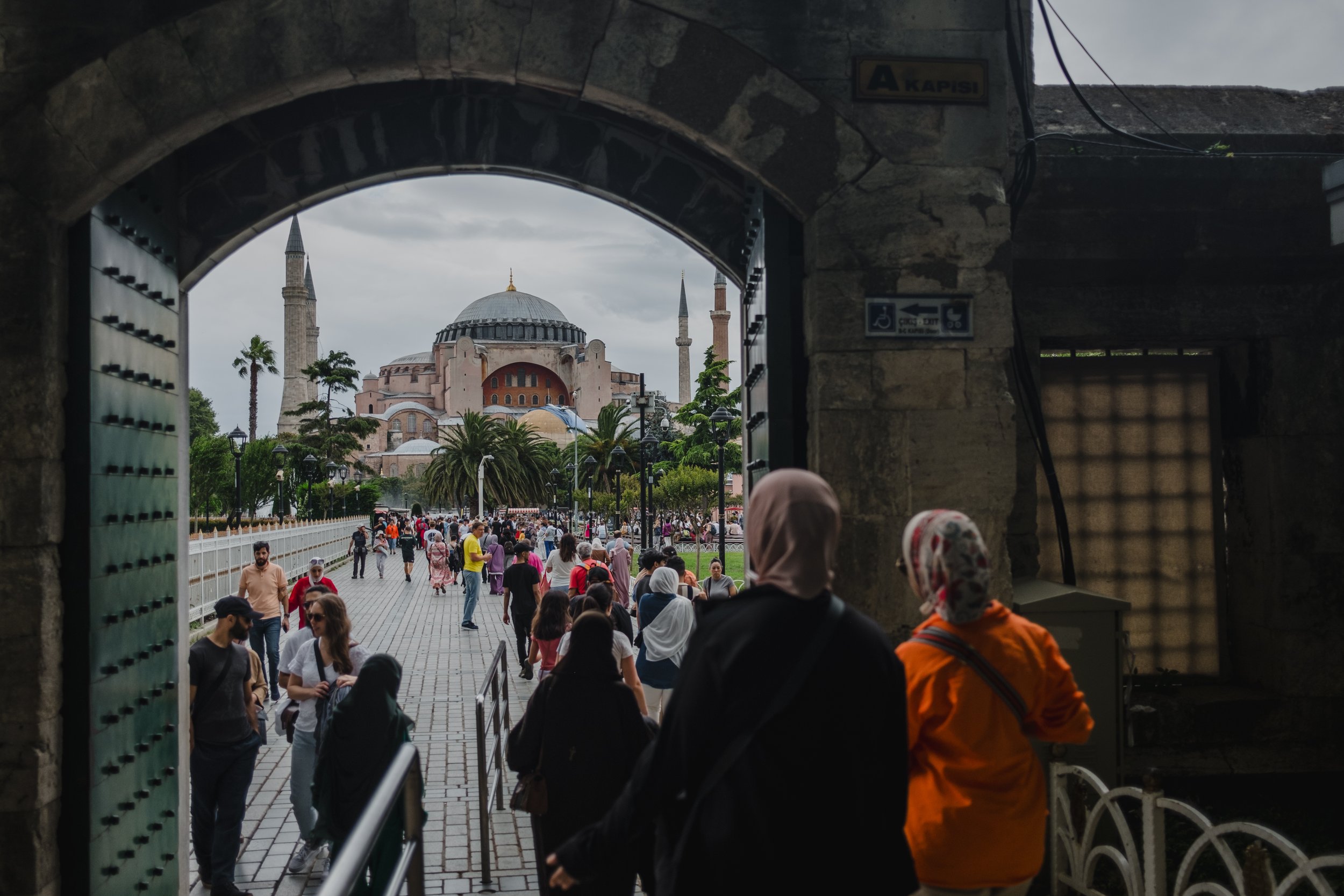 View of Hagia Sophia through an archway, with many people walking in the courtyard, some with hijabs and others in casual clothes, overcast sky in the background.