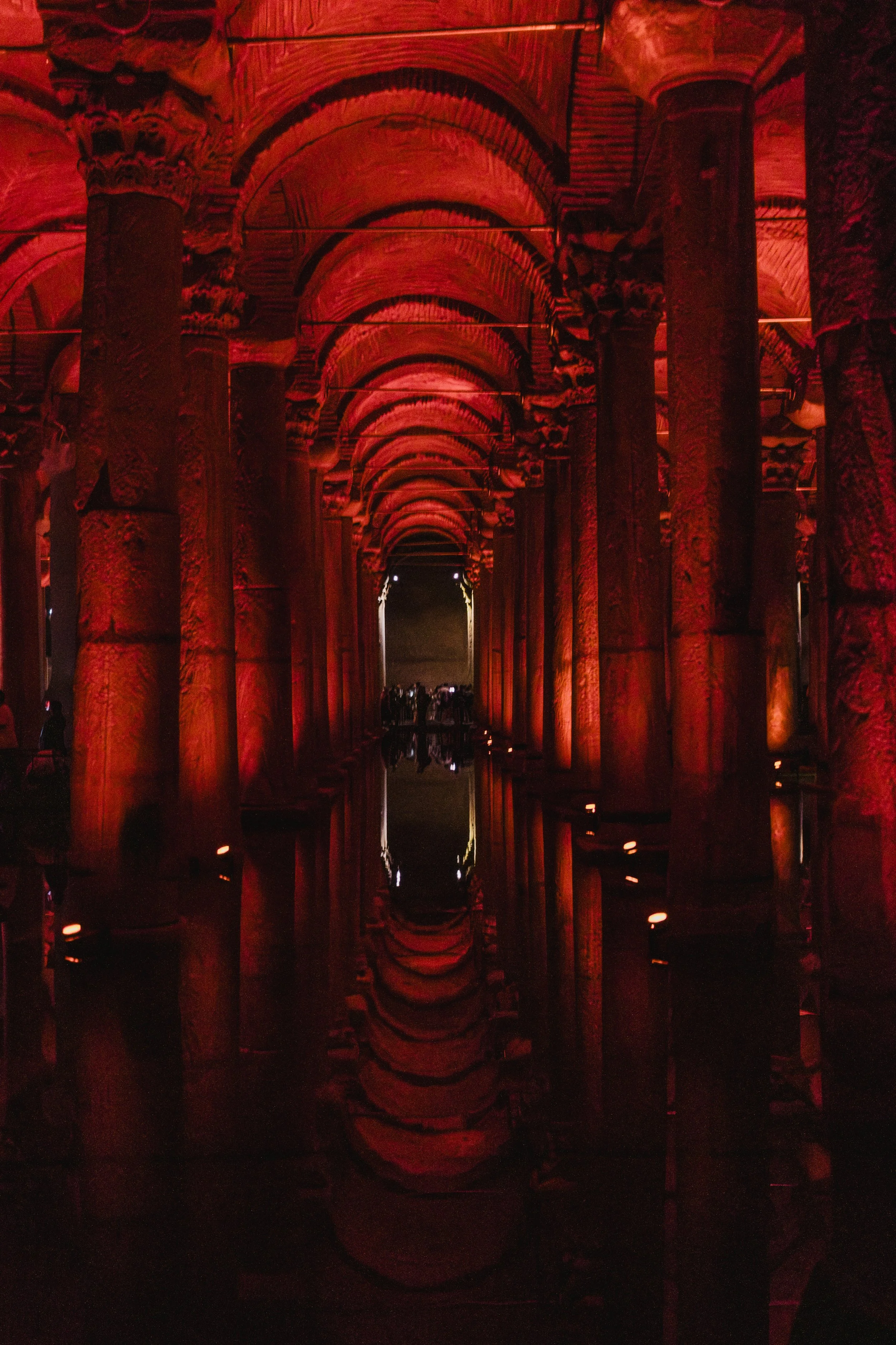 A dimly-lit underground space with columns and arches illuminated in red light, reflecting on a shallow pool of water.