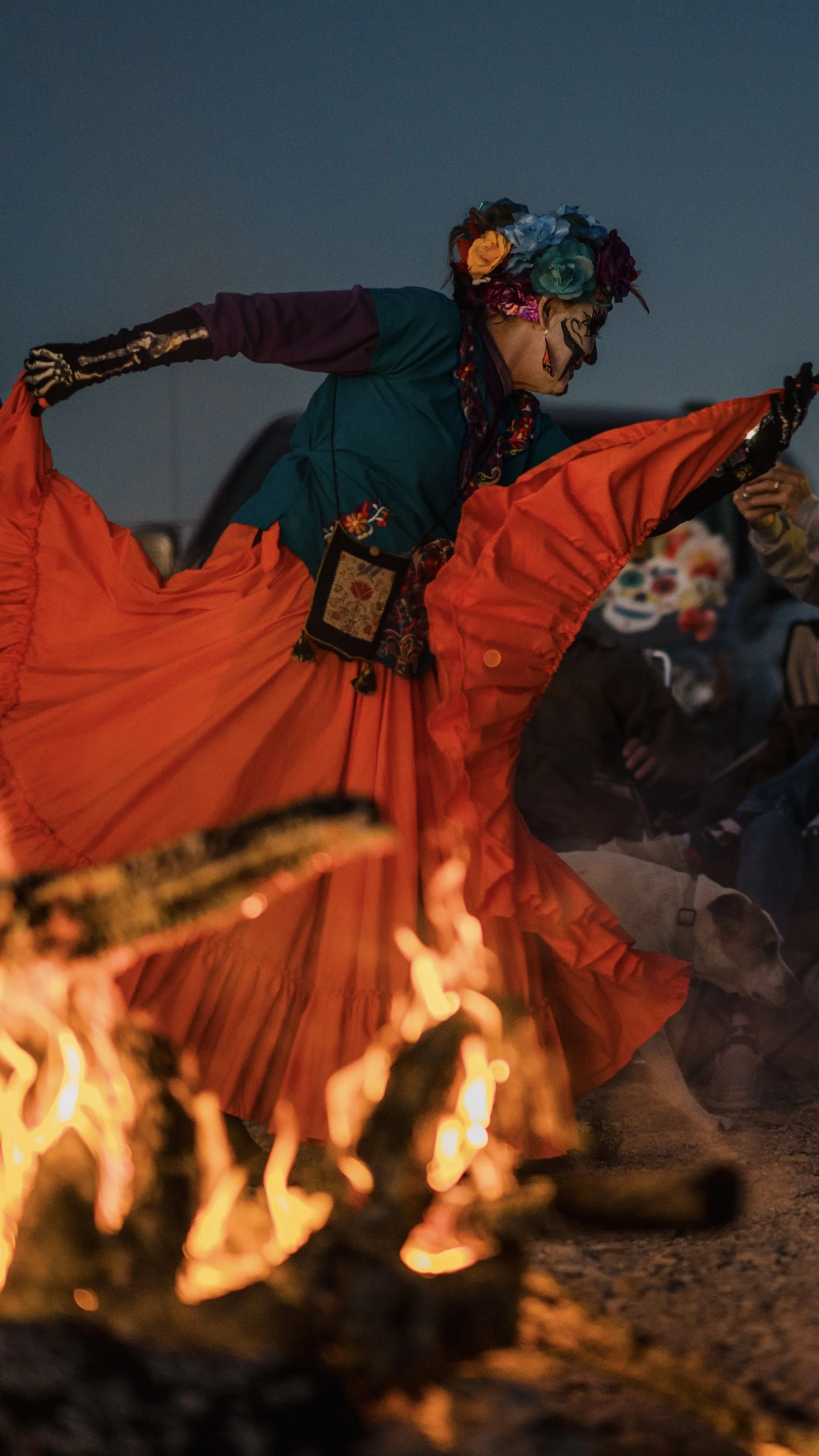 Person dressed in colorful costume, wearing flowers in headwrap and face makeup, dancing near campfire at night.