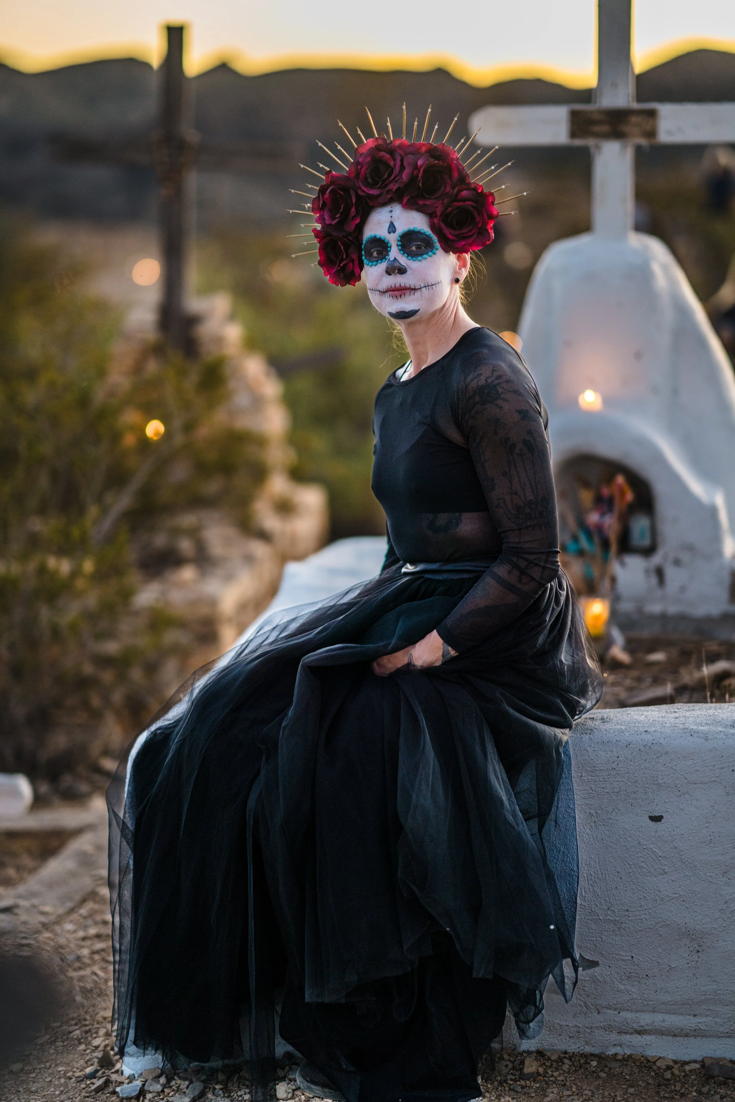 Person dressed in black tulle dress with sugar skull face paint, wearing a headpiece of red roses and gold spikes, sitting outdoors at sunset.