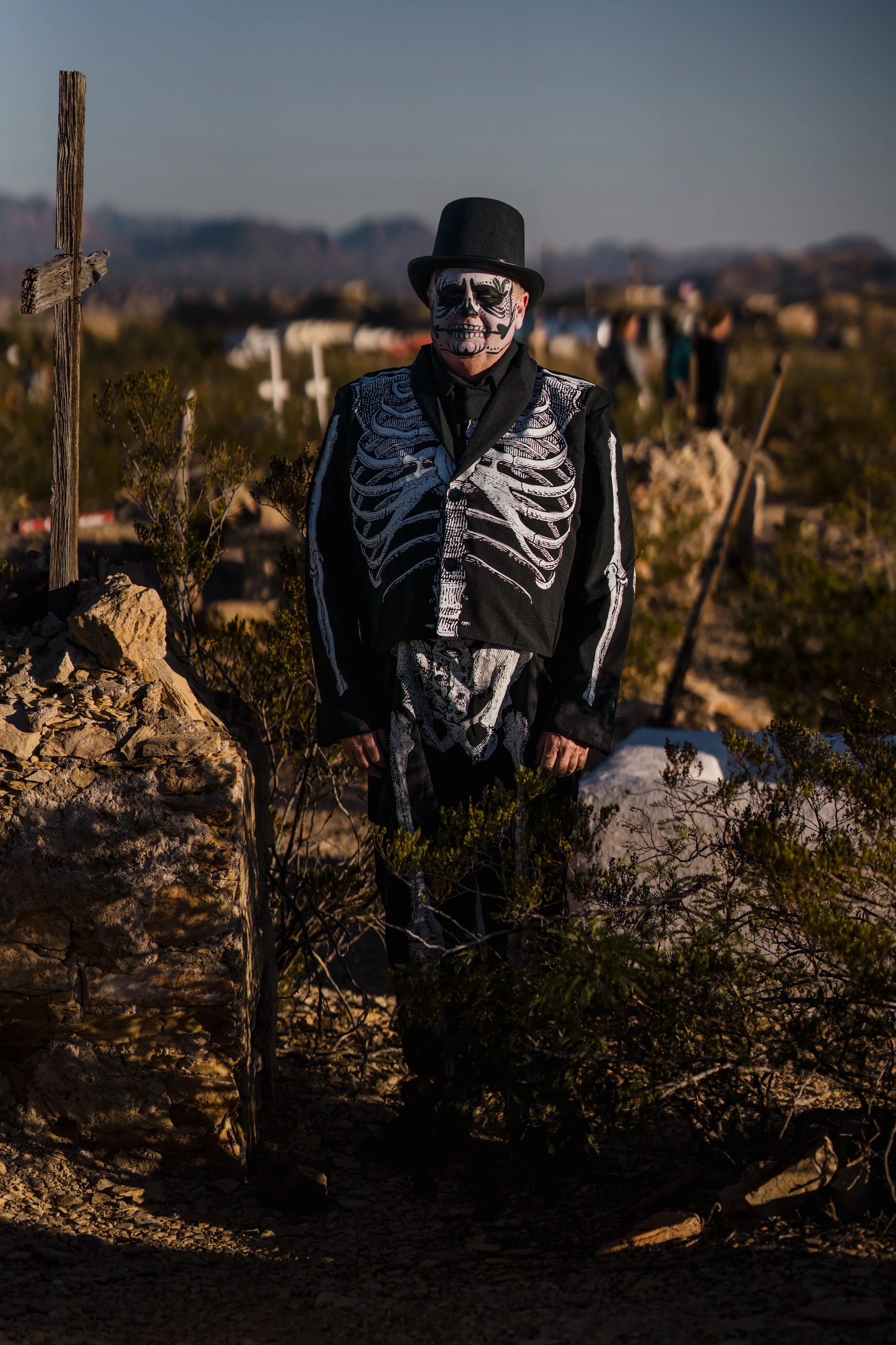 Person dressed as a skeleton in a skull-makeup costume, wearing a black top hat and black clothing with white skeleton print, standing in a desert landscape during sunset.