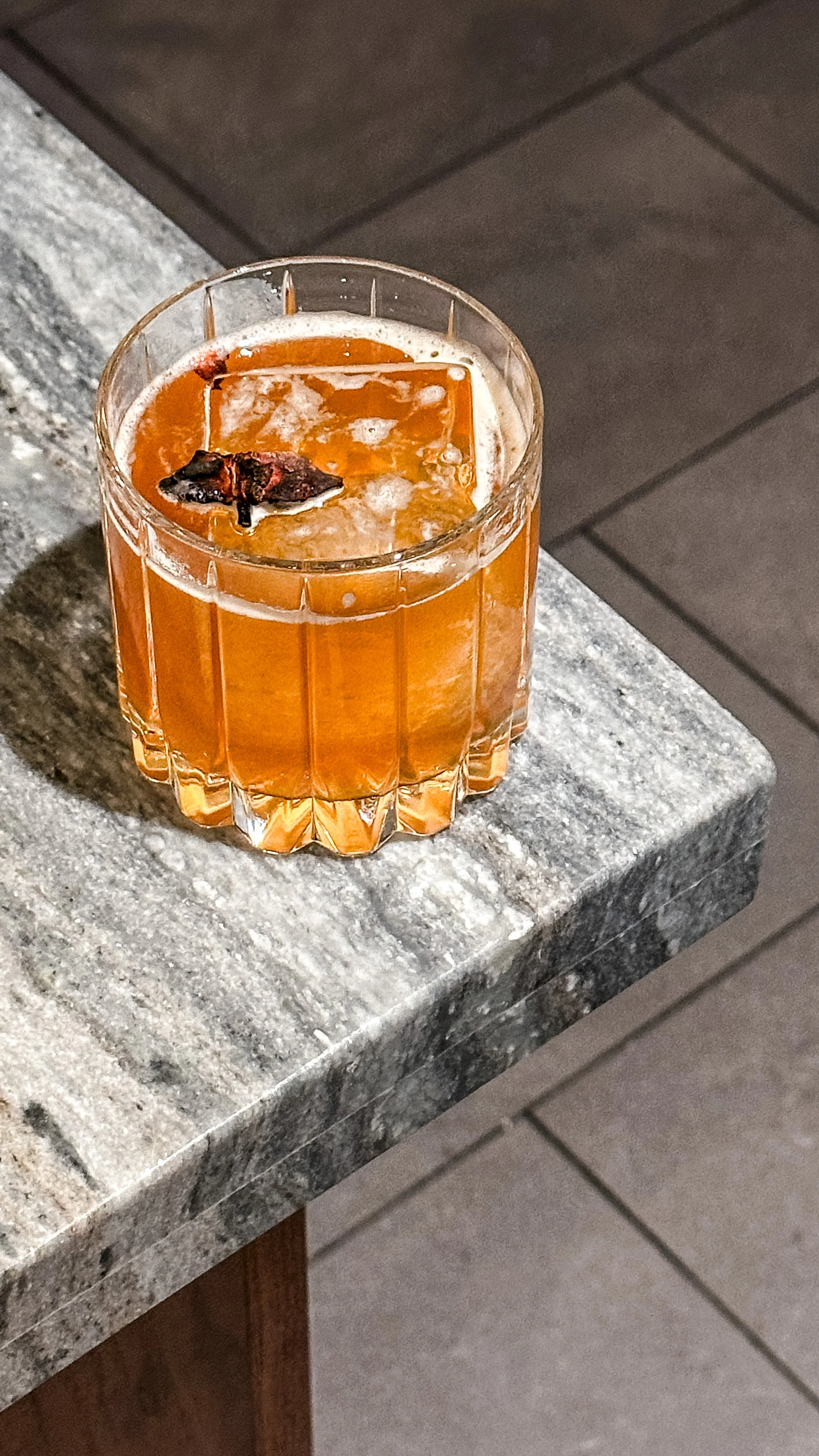 A glass of an amber-colored beverage with ice and a cherry garnishment on top, placed on a gray granite countertop.