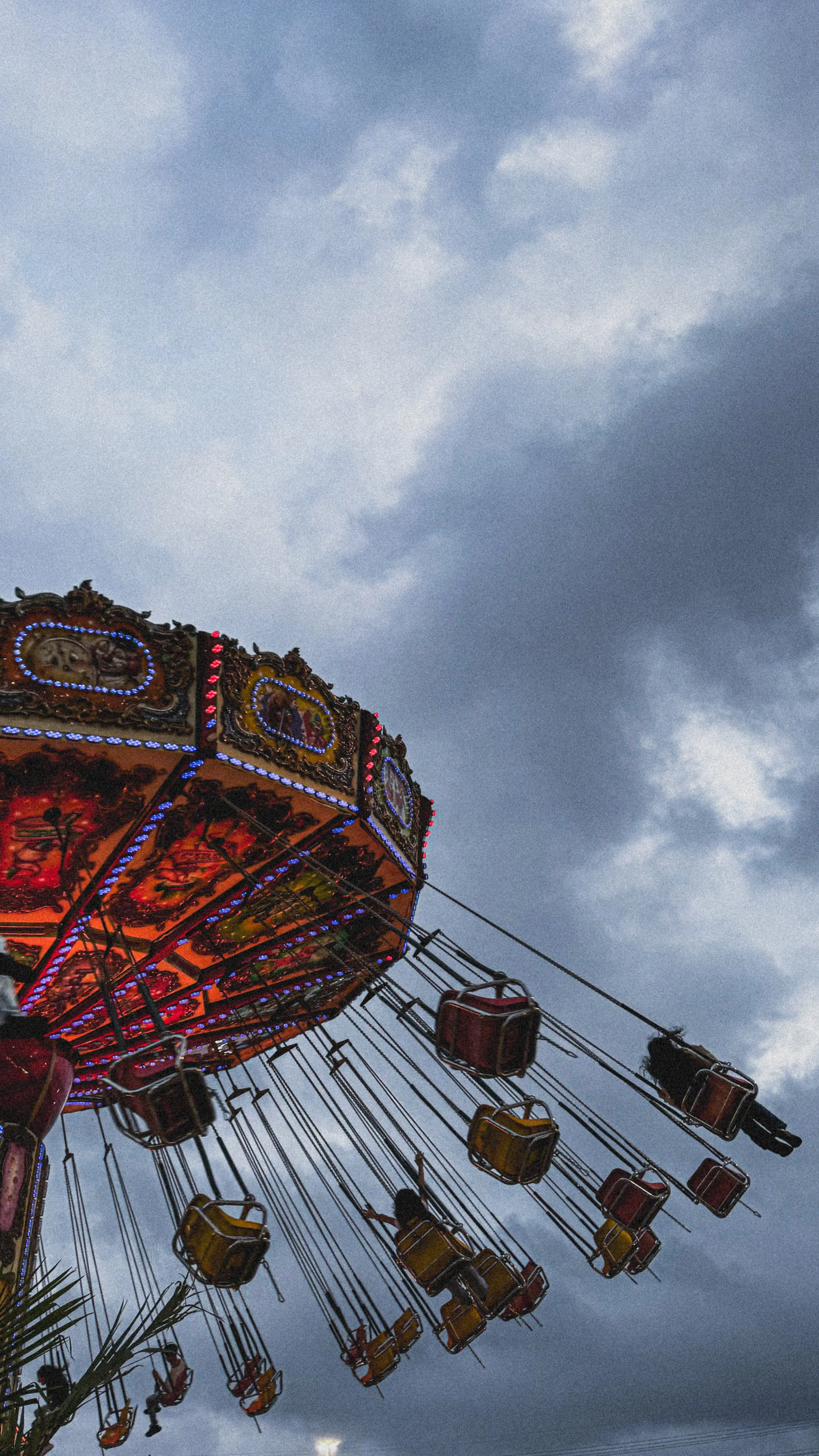 A colorful swing ride at a fair with blue, red, and yellow swings in motion, set against a cloudy sky in the background.