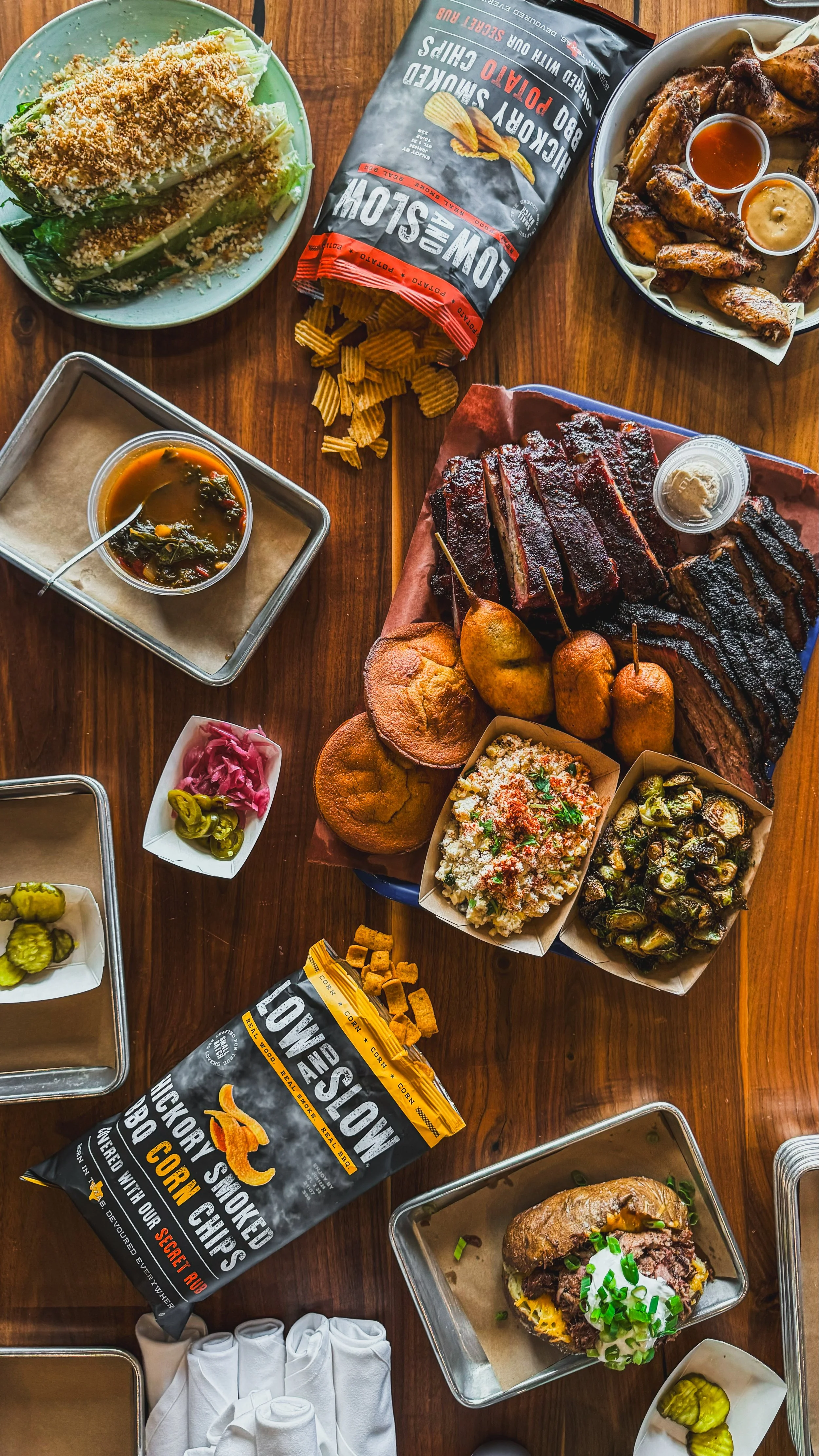 A variety of Southern barbecue foods on a wooden table, including ribs, smoked brisket, fried chicken wings, baked cheese bread, potato salad, roasted Brussels sprouts, pickles, pickle chips, and a sandwich with beef, with bags of smoked corn chips a
