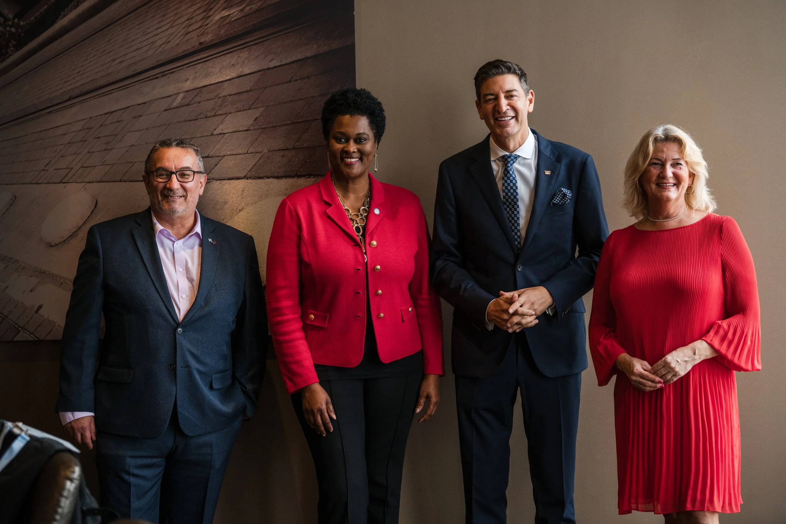 Group of four diverse professionals standing indoors, posing for a photo, two men and two women, dressed in business attire, smiling at the camera.