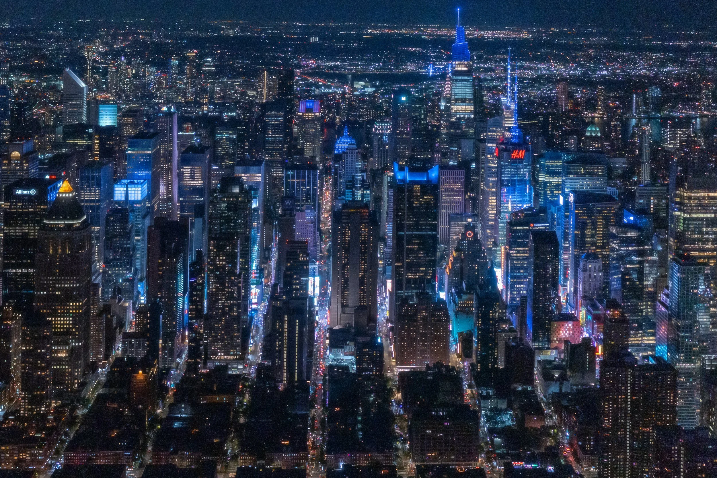 Nighttime aerial view of a city skyline with illuminated skyscrapers and streets, including recognizable buildings with bright neon lights in New York City.