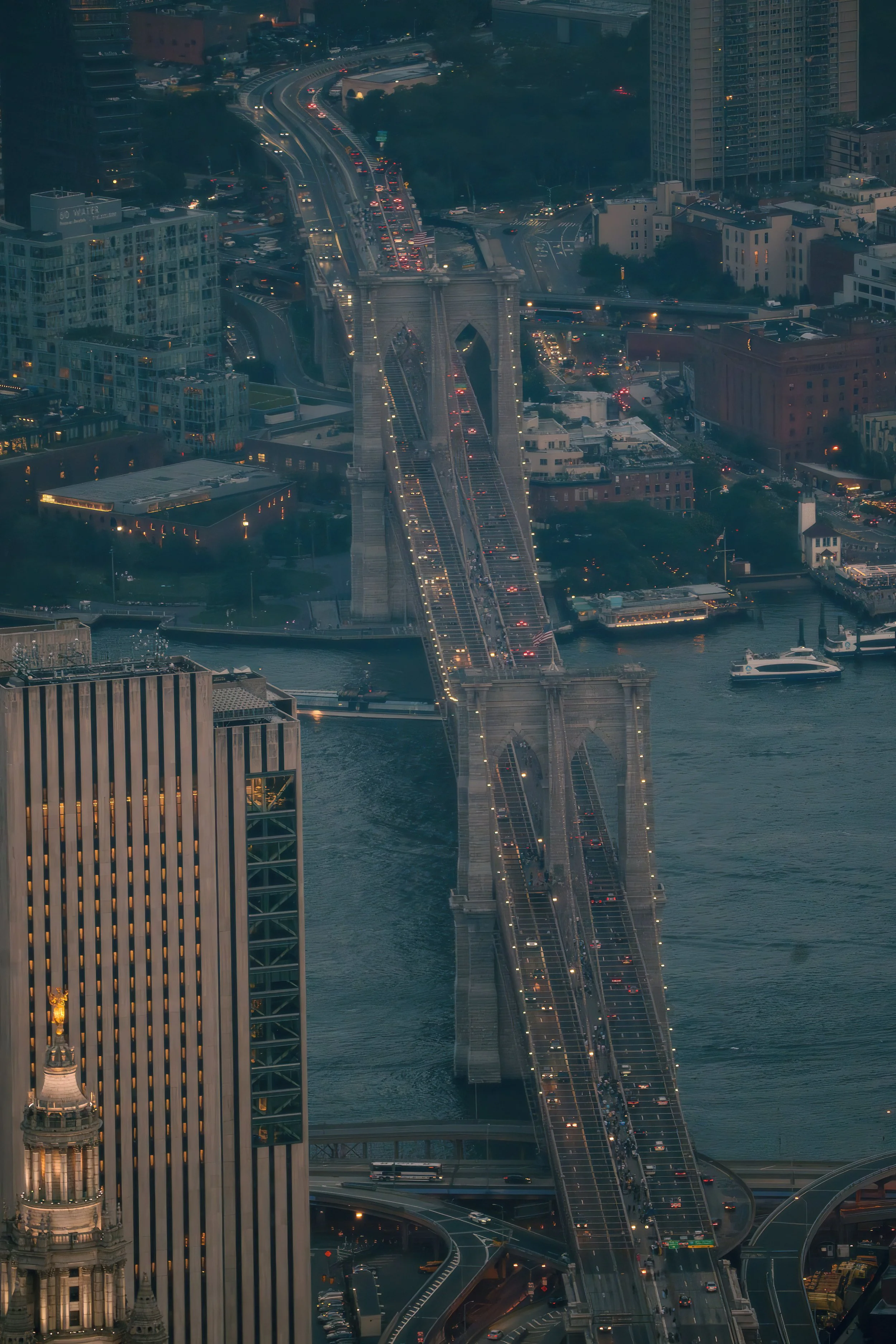 Aerial view of the Brooklyn Bridge in New York City at dusk, with traffic on the bridge and surrounding cityscape, including tall buildings and a river.