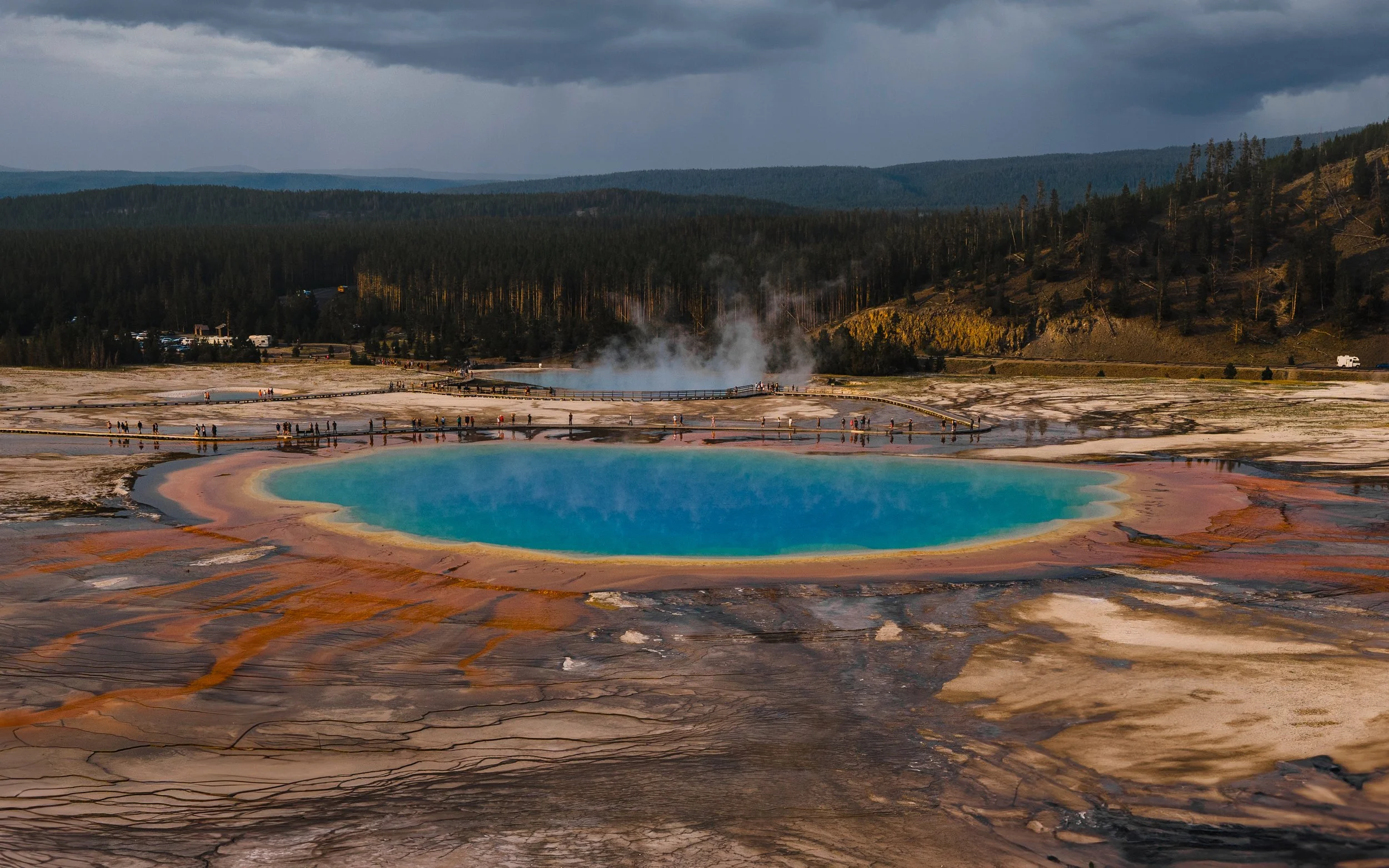 Blue hot spring surrounded by colorful mineral deposits with steam rising in the background, in a geothermal landscape.
