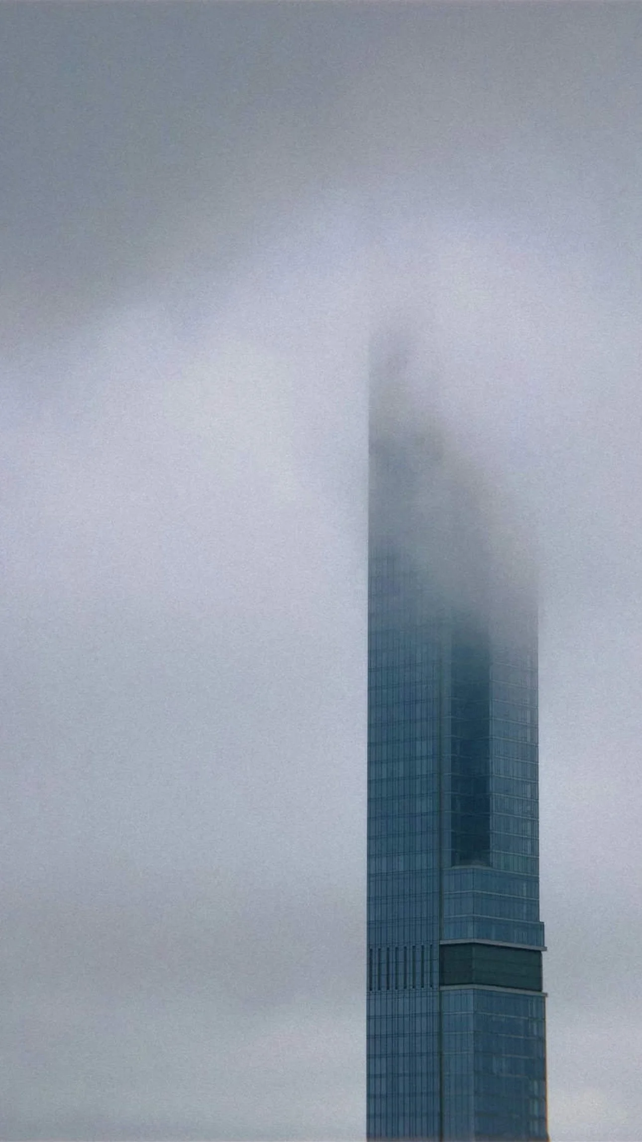 A tall skyscraper with glass windows, partially obscured by fog or low-hanging clouds.