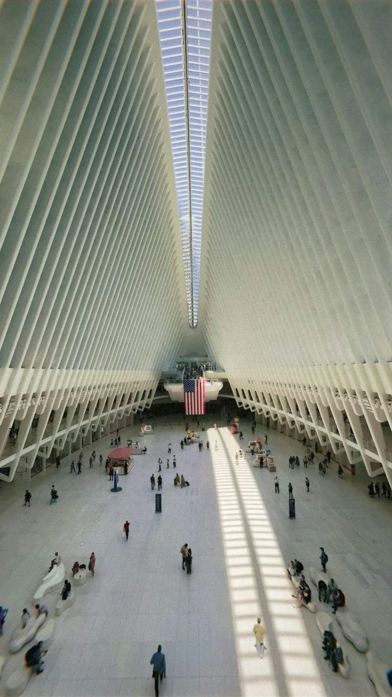 Interior view of the Oculus at the World Trade Center, featuring tall white architectural columns, natural light coming through a skylight, an American flag hanging in the center, and visitors walking and sitting inside.