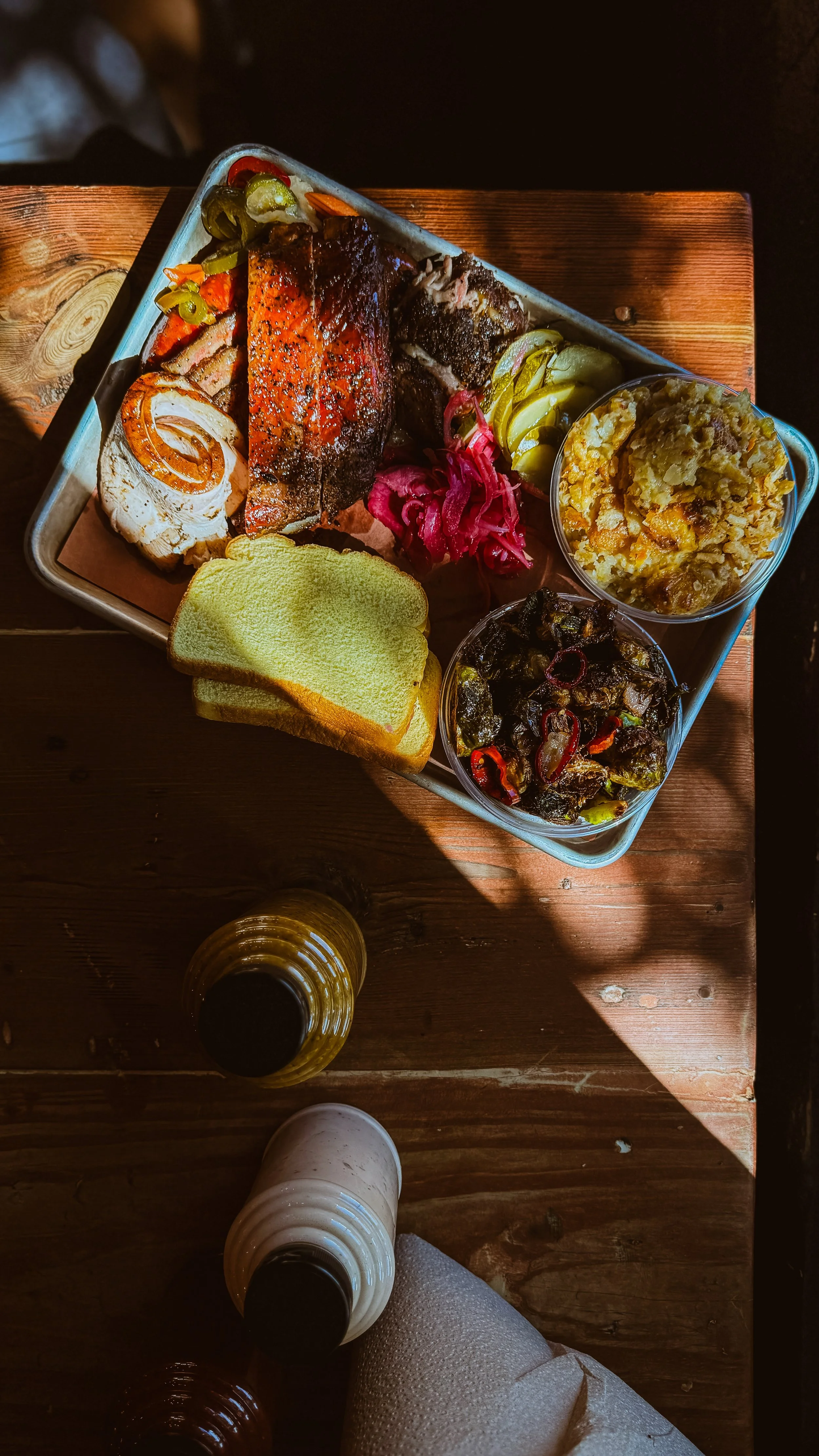 A metal tray filled with grilled fish, bread, pickled vegetables, mashed potatoes, and cooked greens, served on a wooden table with glasses of beer nearby.