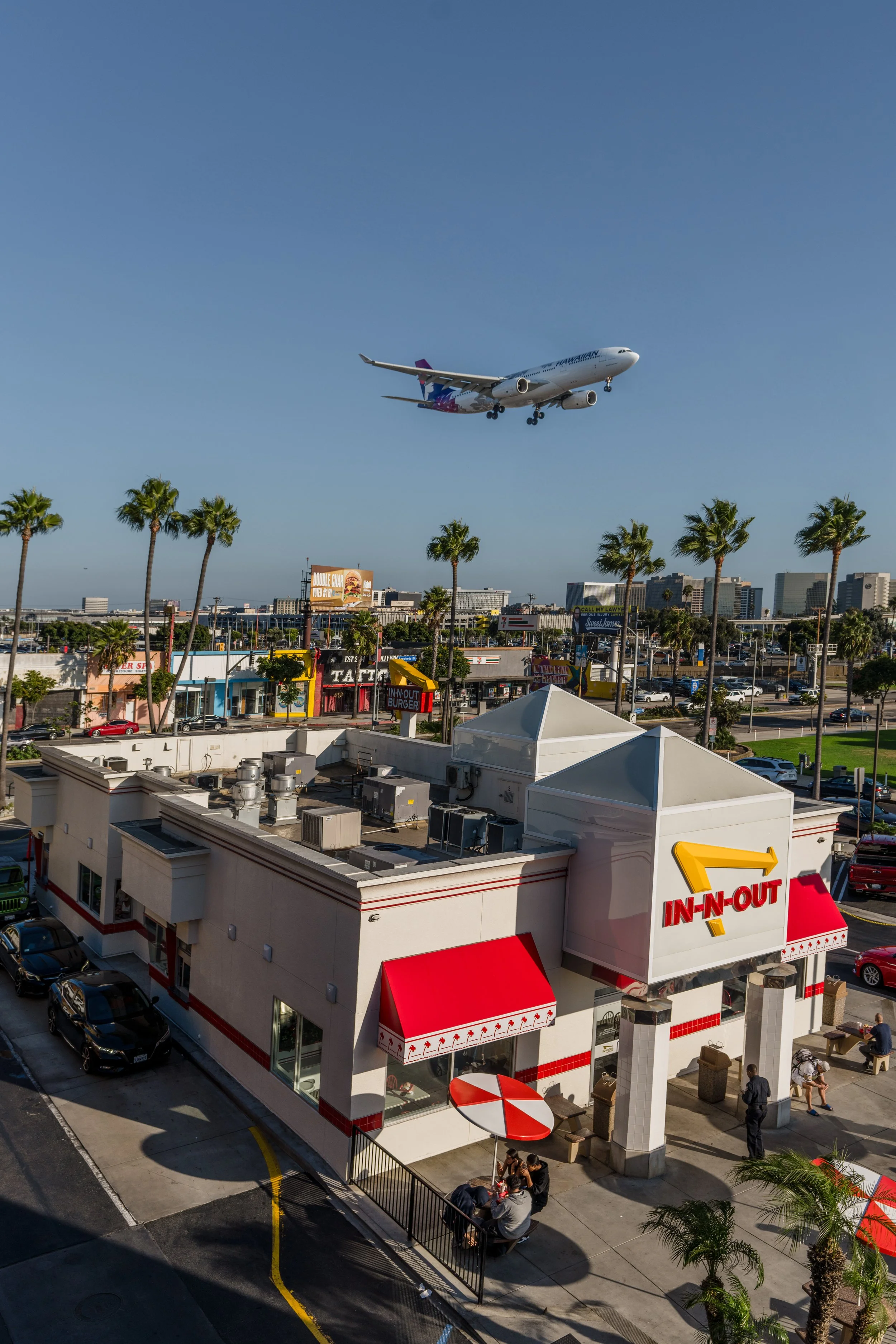 An airplane flying low over an In-N-Out Burger restaurant, with palm trees, vehicles, and buildings in the background on a sunny day.
