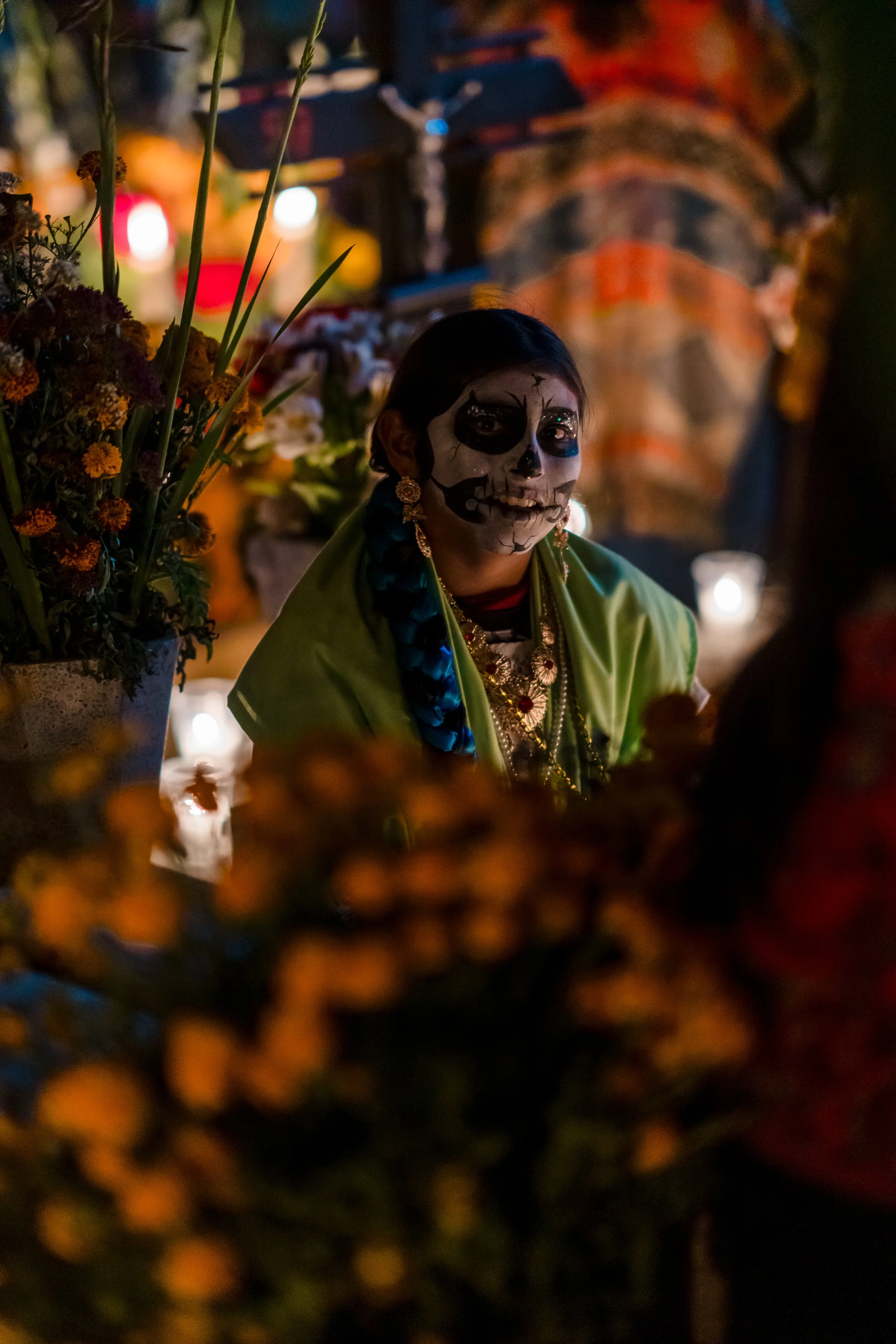 Young girl with face painted as a sugar skull, wearing traditional colorful jewelry and clothing, surrounded by flowers and candles at a Day of the Dead celebration.