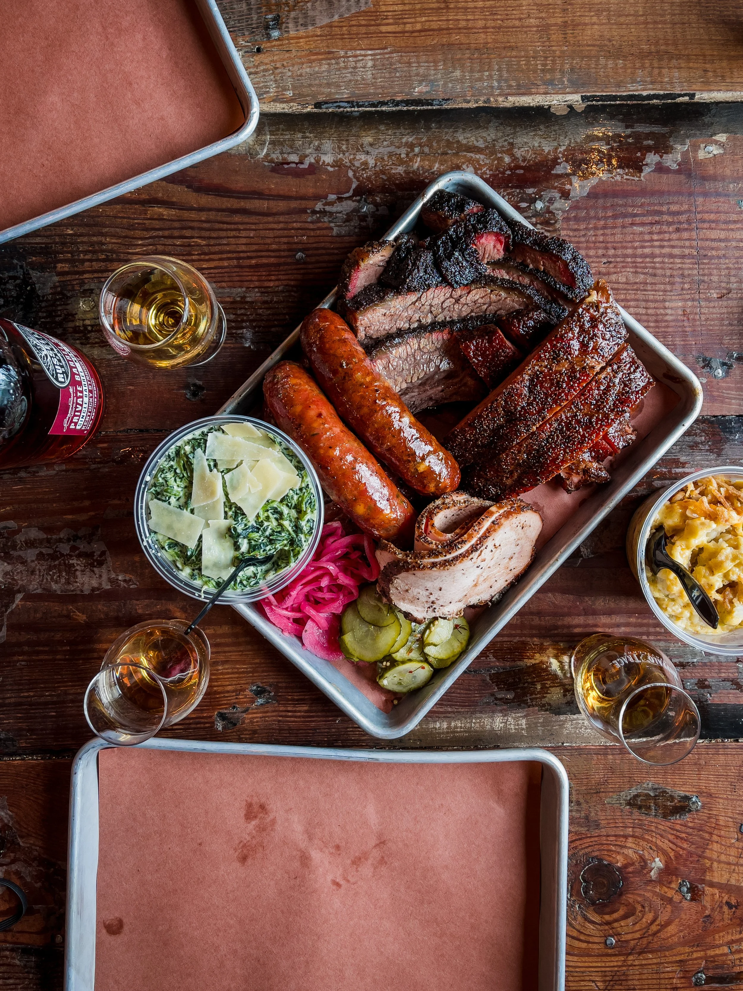 A tray of assorted barbecued meats, including brisket, sausages, ribs, and pulled pork, served with pickles and onions, topped with sides of creamy spinach with cheese and mashed potatoes, and glasses of beer on a rustic wooden table.